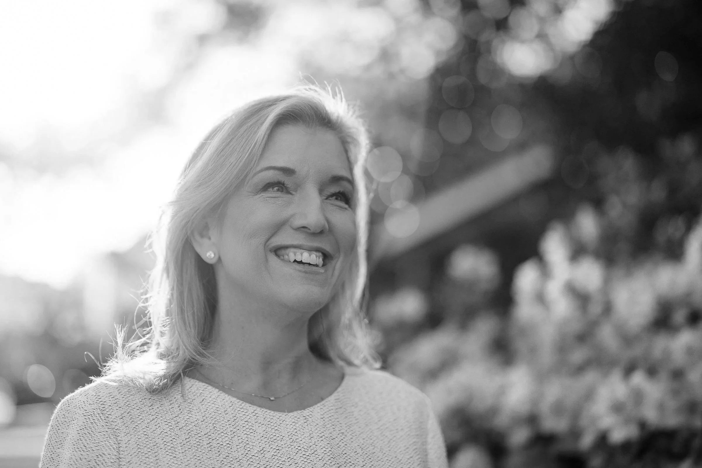 A woman with long hair and earrings smiling outdoors with blurred trees and sunlight in the background.