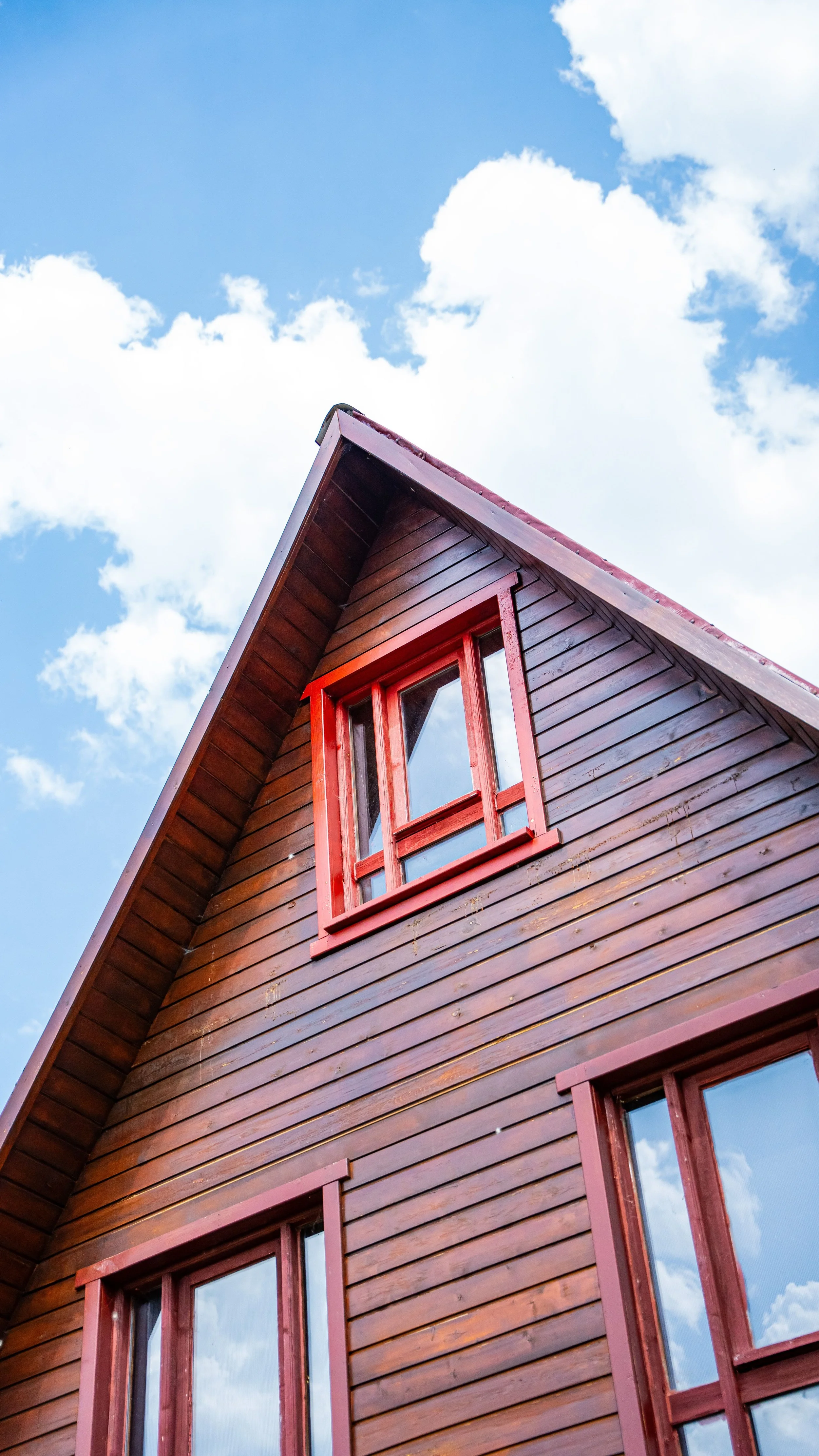A wooden house with red window frames and a steeply pitched roof against a blue sky with white clouds.