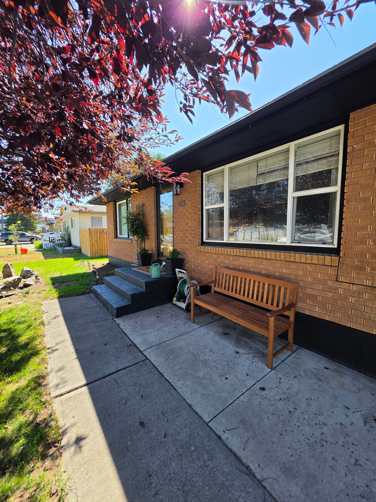 A house with a brick exterior and large windows under a black roof, with a small front steps and a wooden bench outside. There are potted plants on the steps, a garden hose, and a yard with grass and a tree with dark red leaves. The sky is clear and blue, with the sun shining.