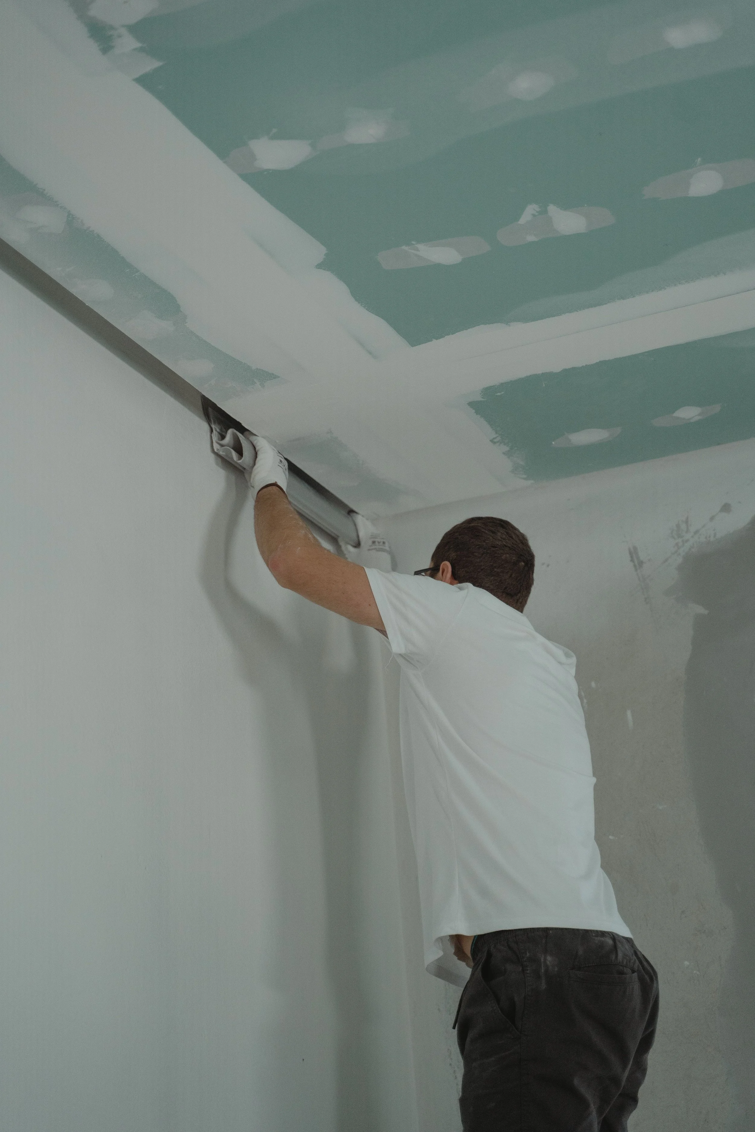 Person installing drywall on ceiling, painting, or working on drywall joint tape, wearing a white t-shirt and gloves.