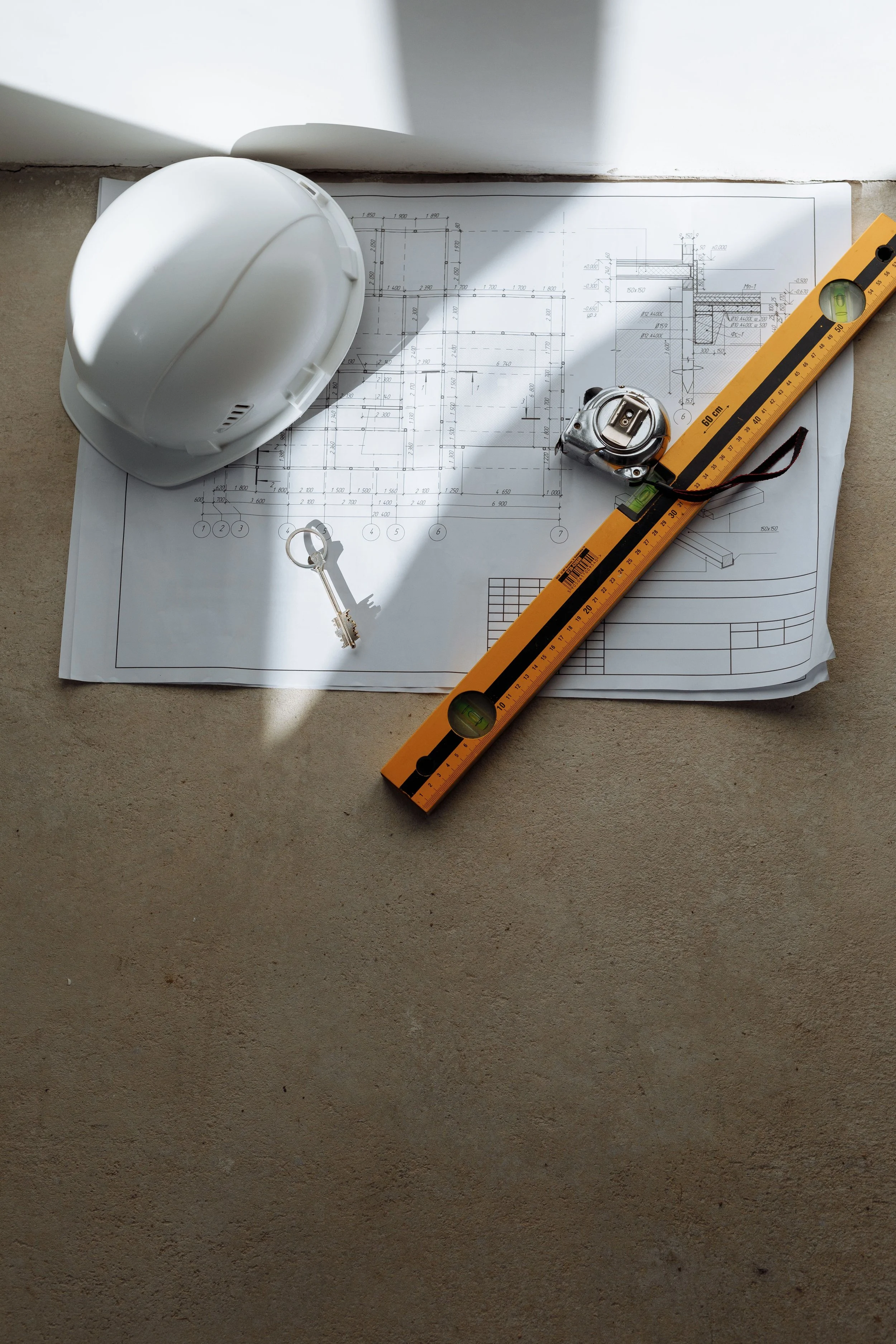 Construction helmet, measuring tape, key, blueprint, and level on a concrete floor.