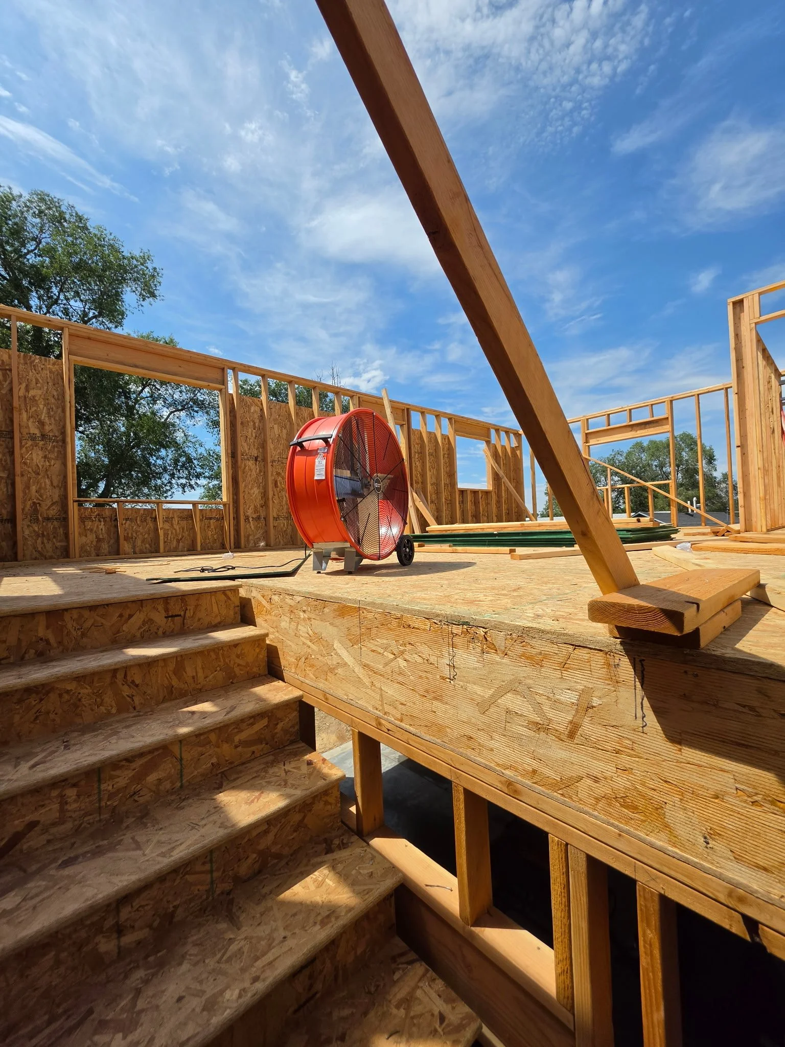 The image shows a construction site of a wooden house with exposed framework. Wooden stairs lead up to the second floor. A red industrial fan is positioned on the floor, and construction materials are scattered throughout. The sky is blue with wispy clouds, and trees are visible in the background.