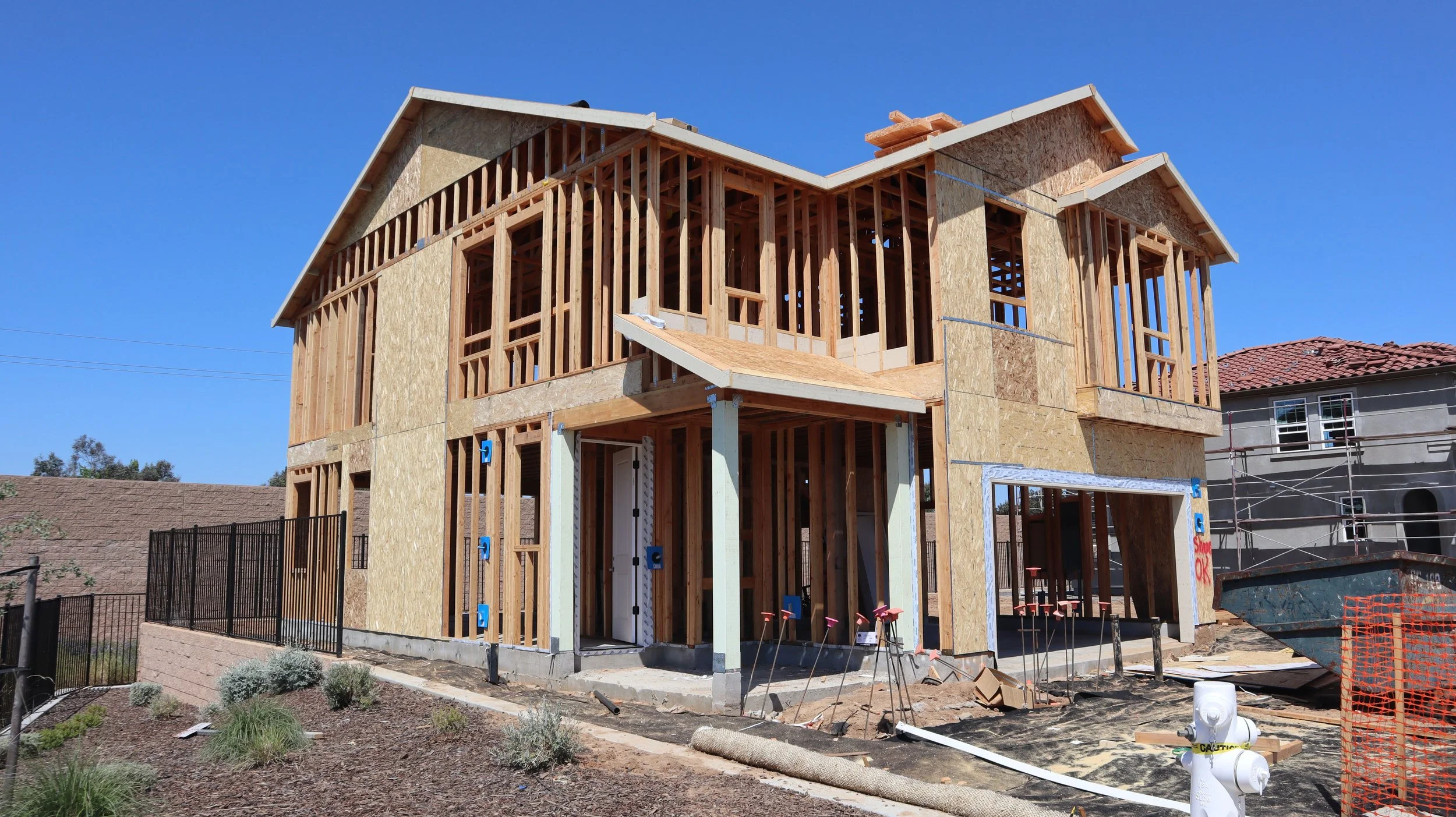 A house under construction showing a wooden frame and partially installed walls with plywood sheathing, set against a clear blue sky.