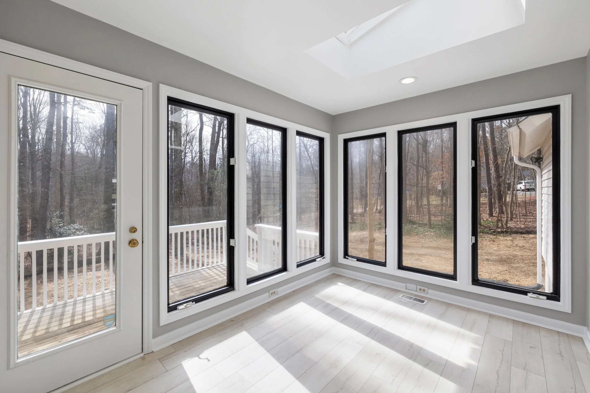 A sunroom with large windows and a door leading to a wooden deck outside, overlooking a wooded backyard.
