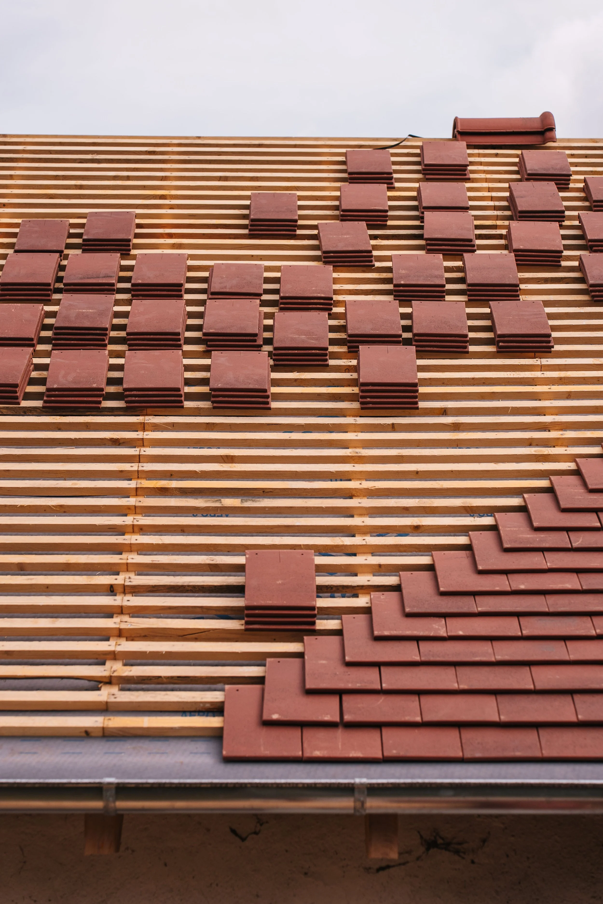An under-construction building roof with wooden batten strips and several red roofing tiles stacked in organized piles.