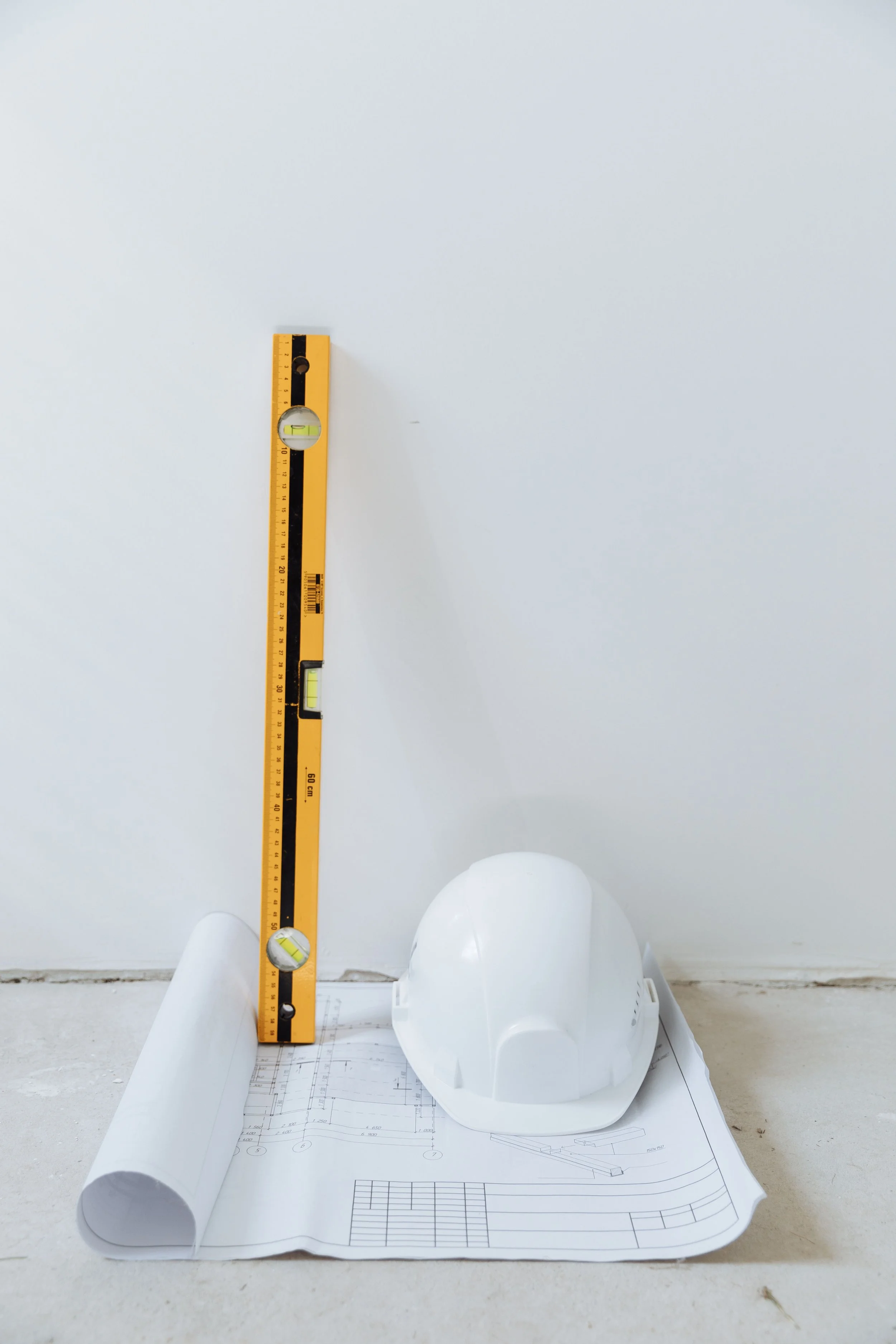 Construction helmet, blueprints, and a yellow measuring tool on a white surface against a white wall.