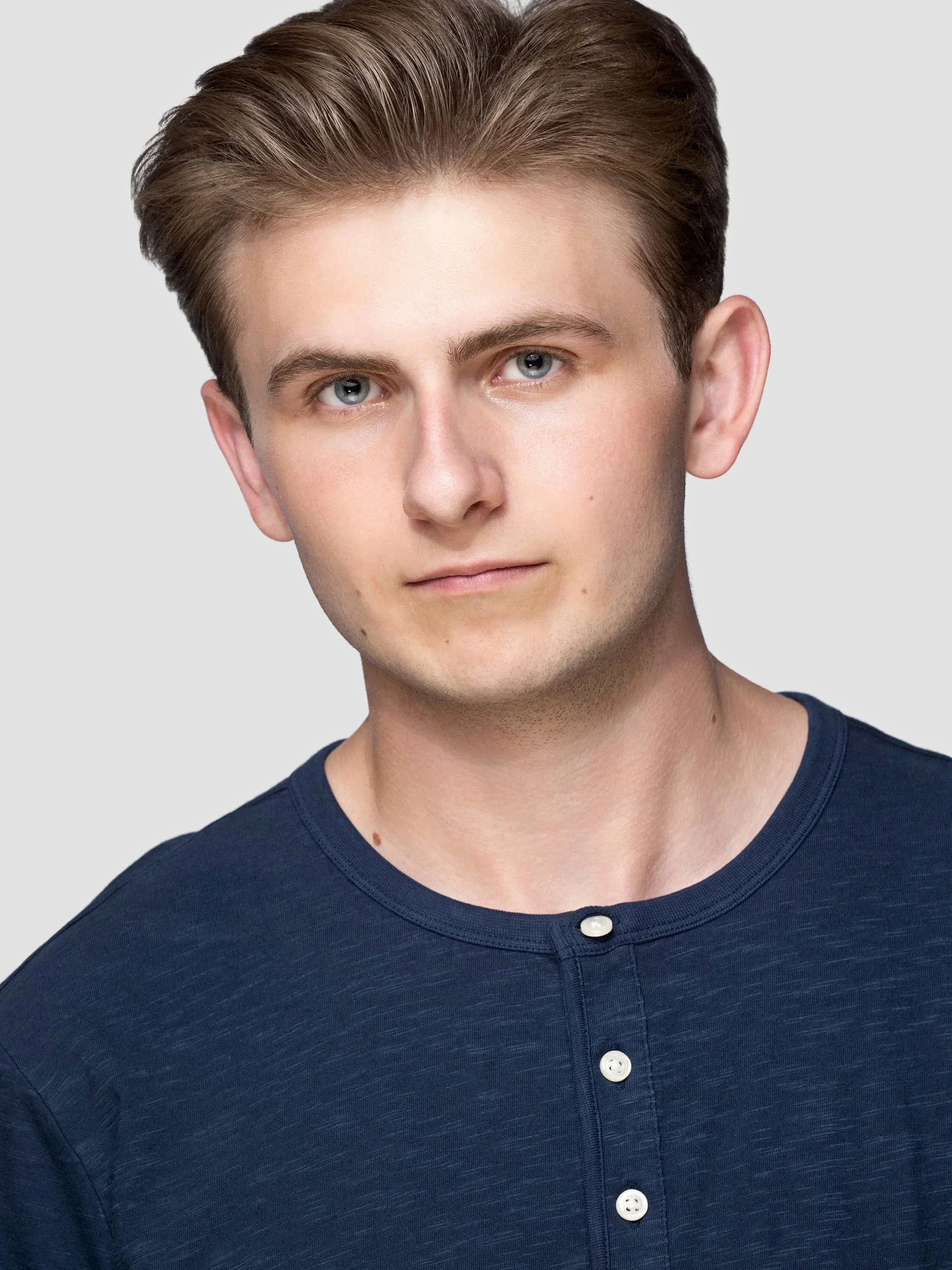 Close-up of a young man with light skin and blue eyes, wearing a navy blue button-up shirt with white buttons, looking directly at the camera against a plain light gray background.