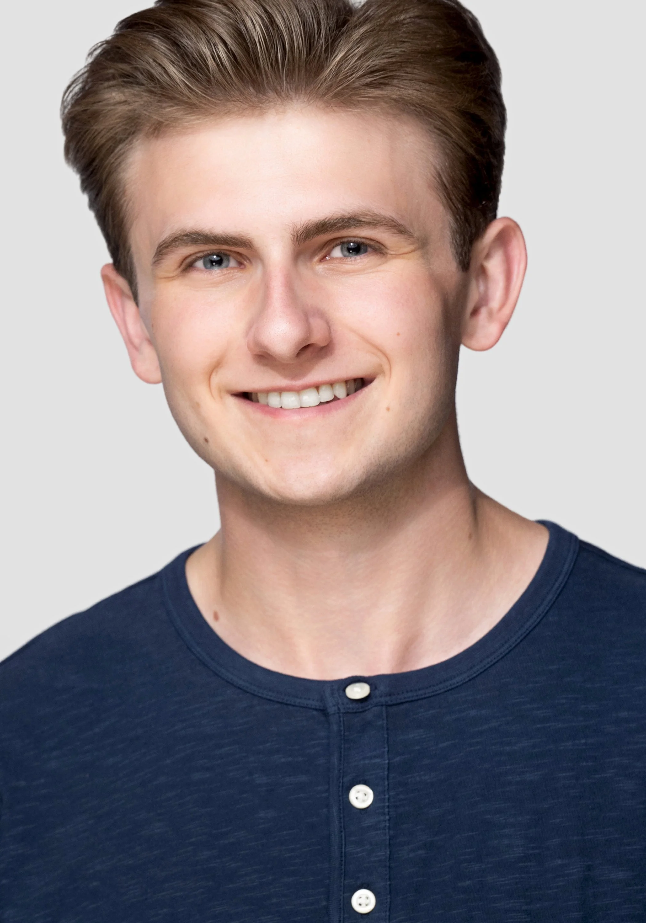 Close-up of a young man with light brown hair, blue eyes, smiling, wearing a navy blue button-up shirt against a plain gray background.