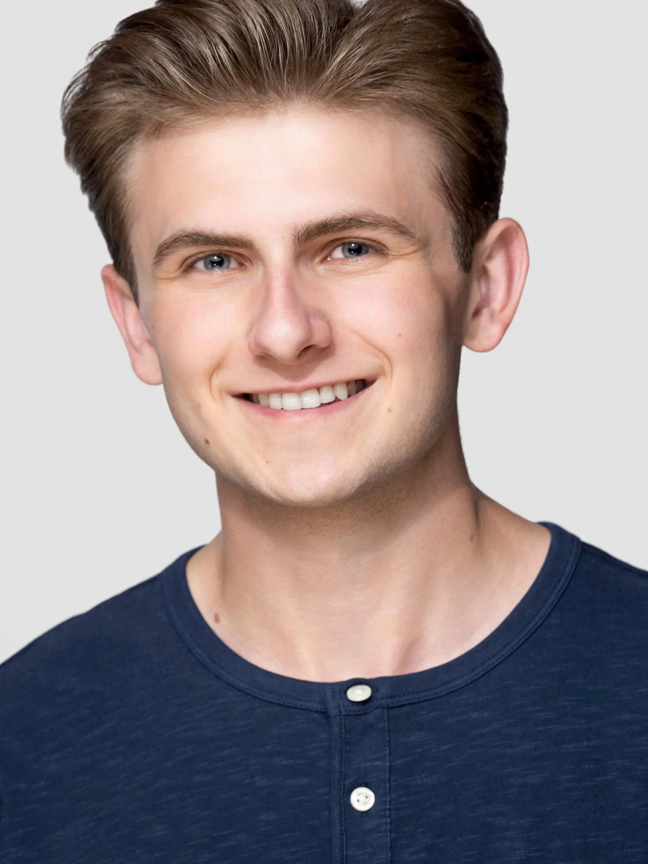 Close-up of a young man with neatly styled brown hair, blue eyes, and a smile, wearing a navy blue shirt with white buttons.
