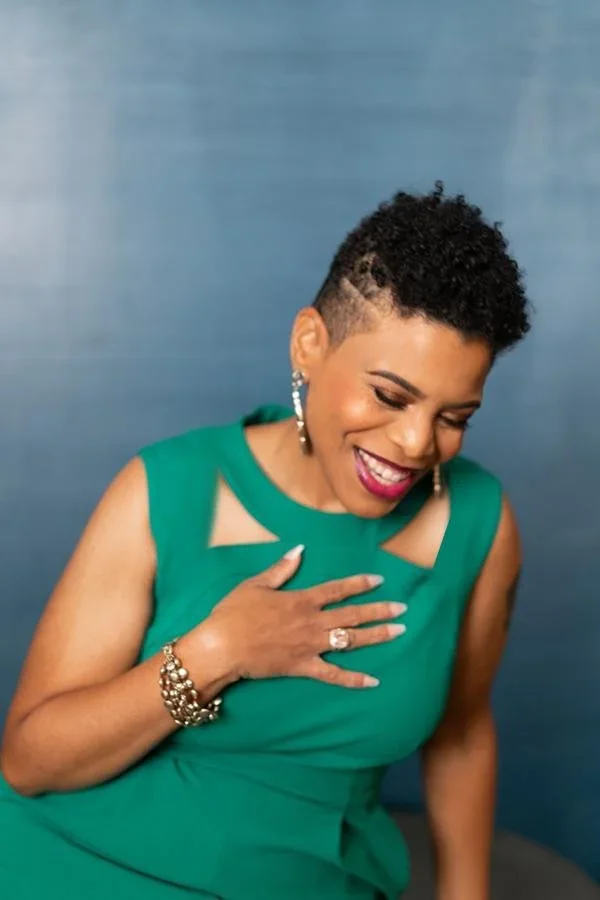 Stacey is pictured with short, curly black hair, wearing a green sleeveless dress, showing a joyful expression with eyes closed and one hand placed on her chest, standing against a blue background.