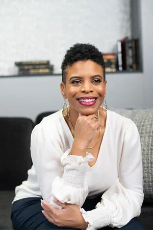 A woman with short curly hair smiling and sitting on a couch in a living room.