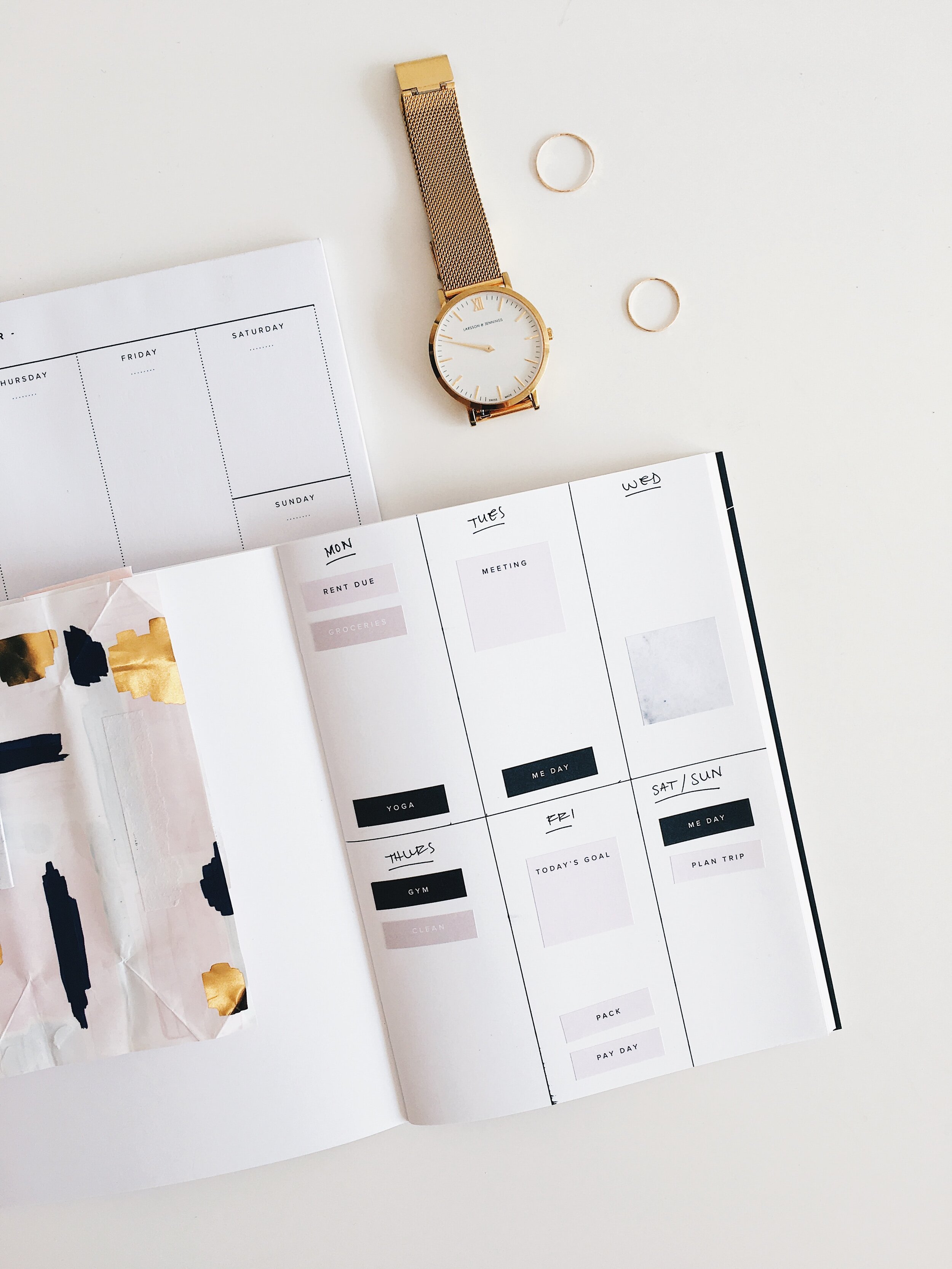 A flat lay of a gold wristwatch with a white face, two gold rings, and a planner or journal with weekly and daily sections on a white surface.