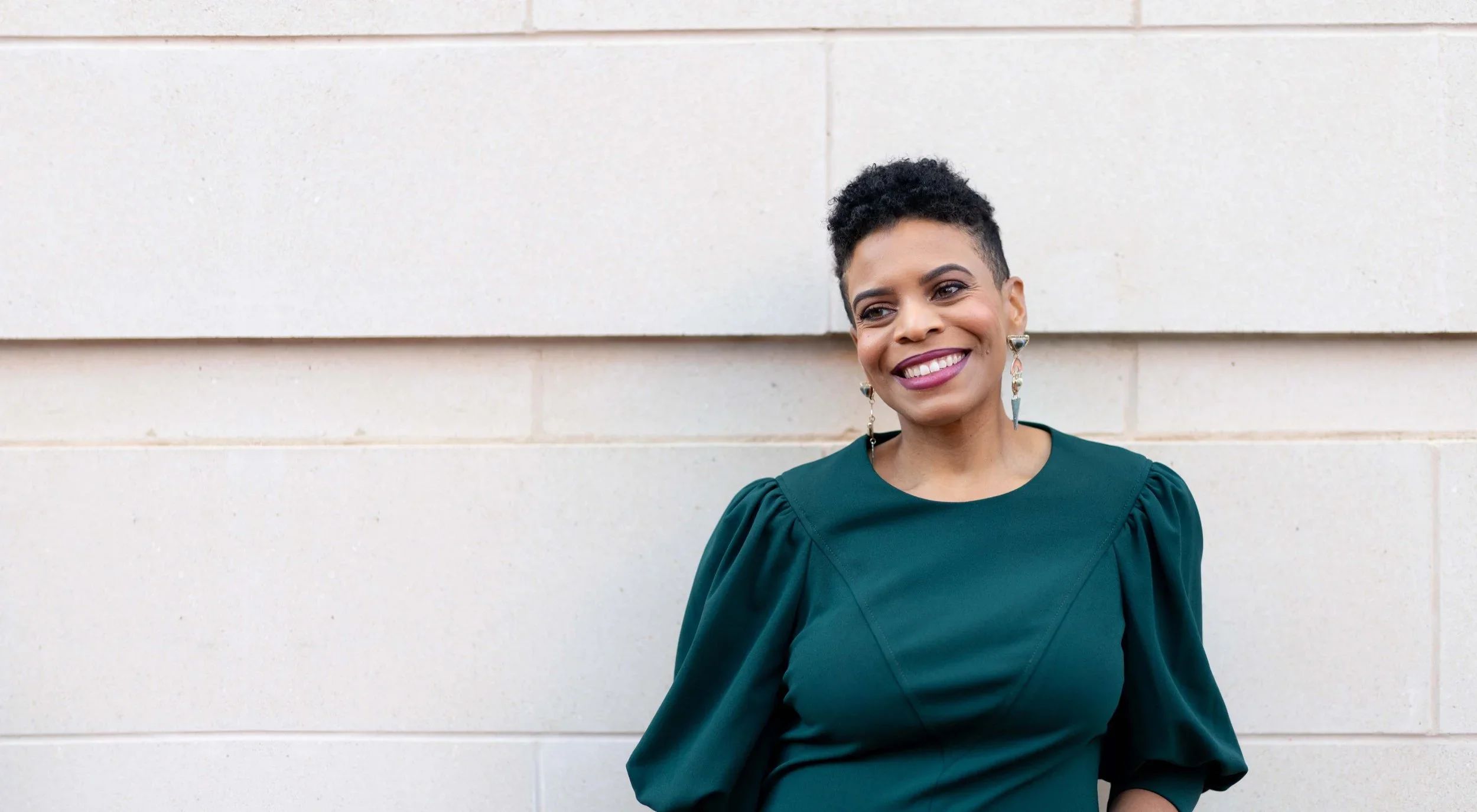 Stacey is smiling with her head tilted to her right (viewer's left). She has dark curly hair styled short, wears a dark green dress and dangling earrings while standing in front of a light-colored wall.