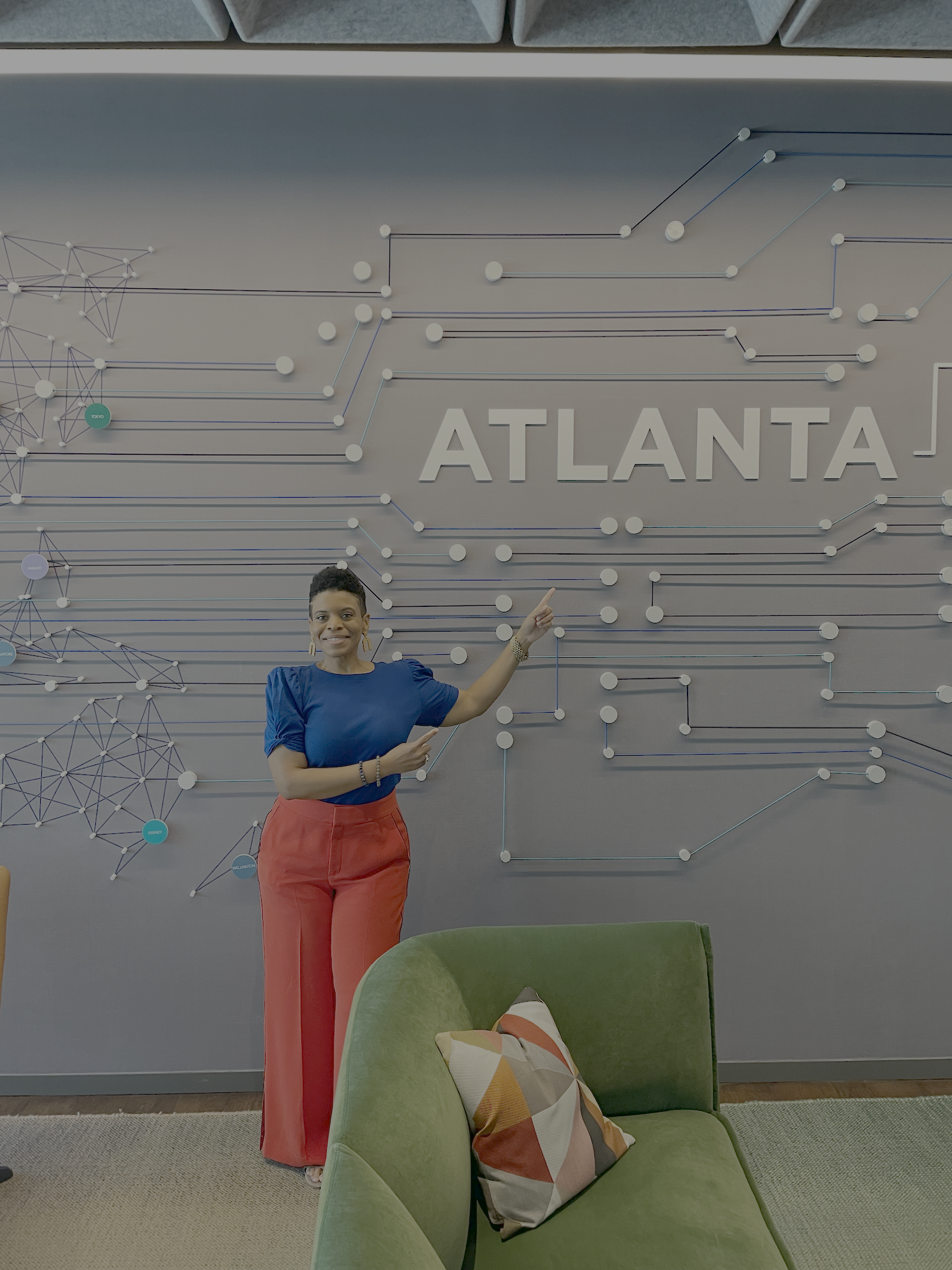 Stacey has short, curly, dark hair wearing a blue top and coral pants, standing in front of a gray wall with the word 'ATLANTA' and a network or map diagram. She is smiling and pointing at the words.
