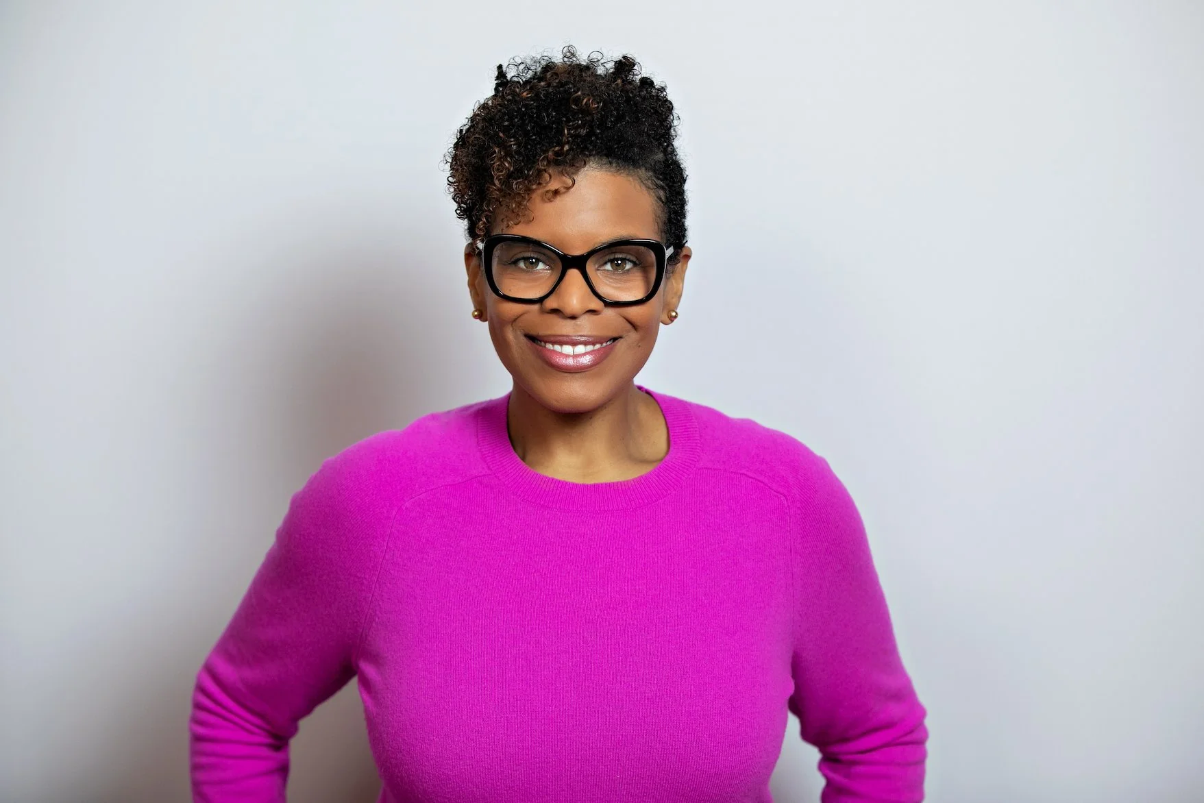 A woman with short curly hair wearing glasses and a bright pink sweater, smiling against a plain light-colored wall.