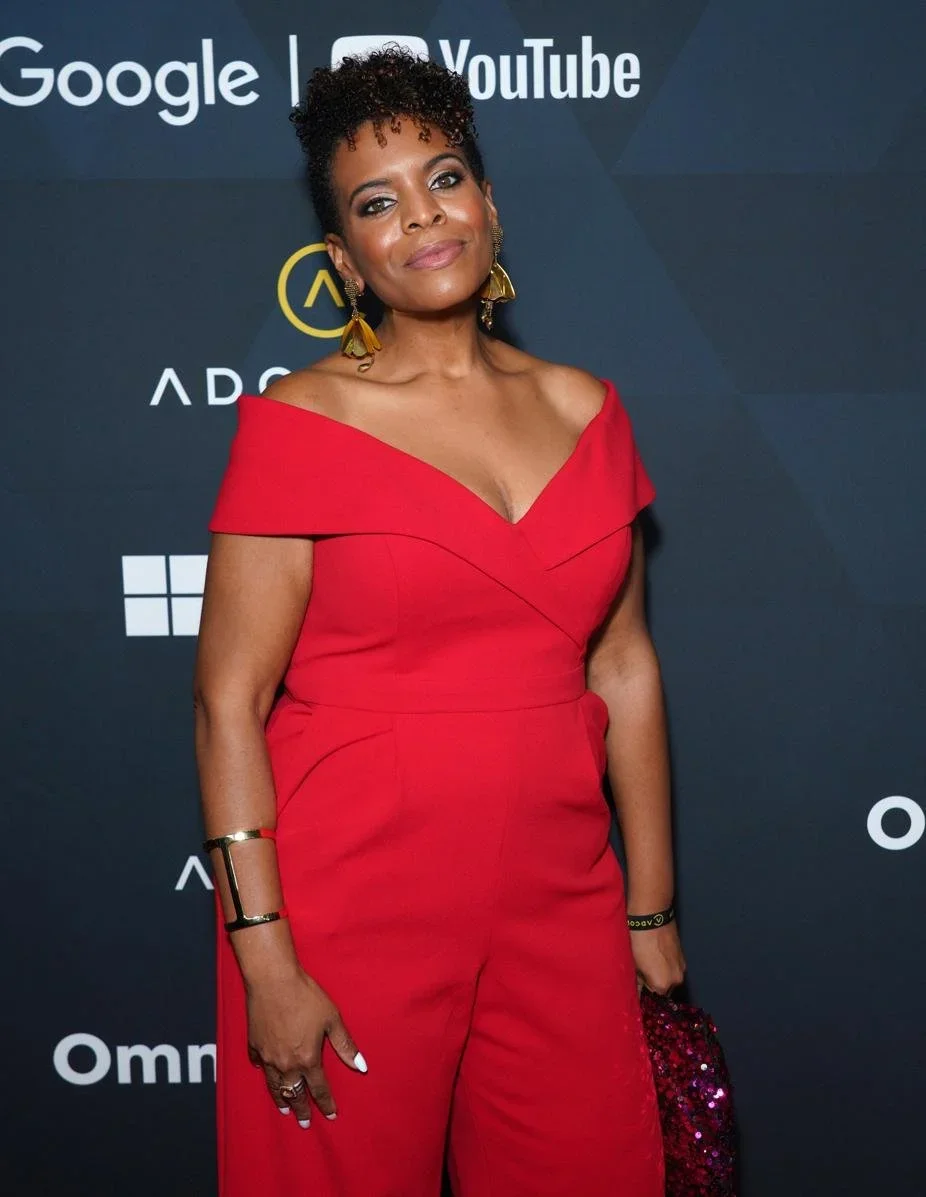 Stacey is pictured on the red carpet at ADCOLOR wearing a red off-shoulder jumpsuit posing on a dark backdrop with logos of conference sponsors on it.