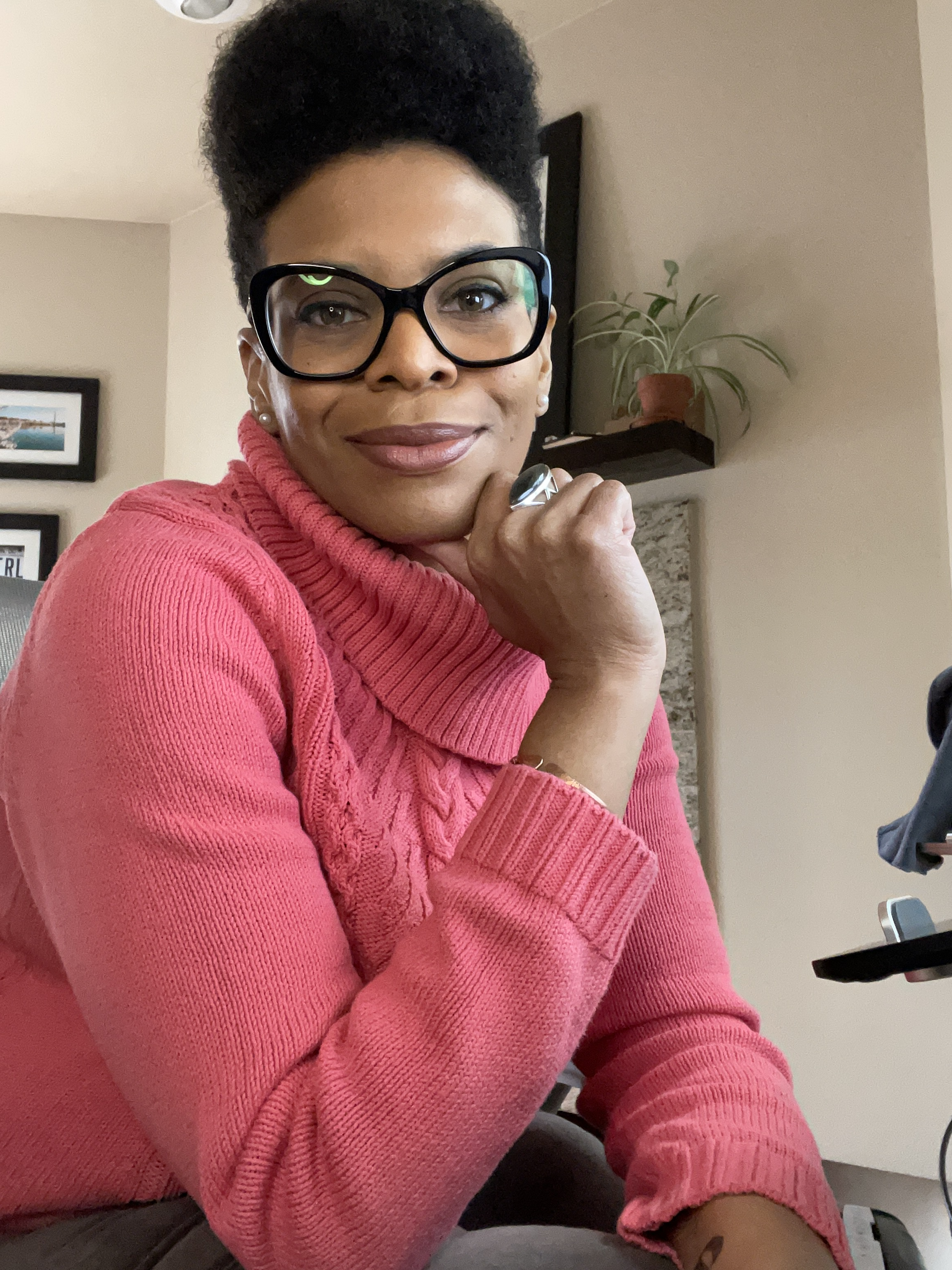 A woman with dark hair styled in a high bun, wearing large black glasses and a pink cable-knit sweater, sitting indoors with a thoughtful expression, resting her chin on her hand, in front of a wall with framed pictures and a shelf with a potted plan