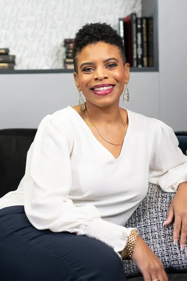 Stacey is pictured smiling with short curly hair, wearing a white blouse, gold earrings, a gold bracelet, and a gold necklace, sitting on a black and white couch in a modern room with a gray wall and a shelf with books in the background.