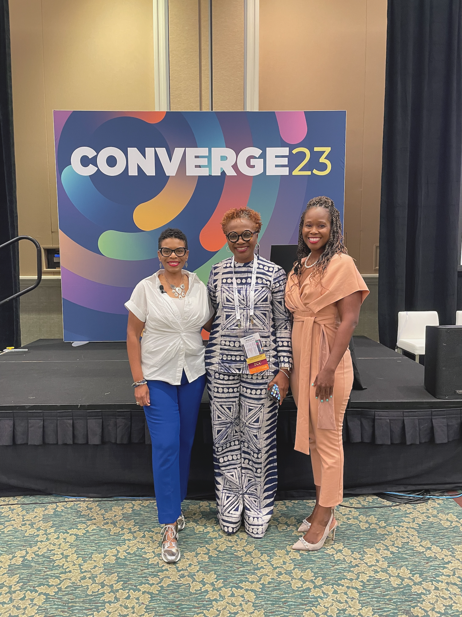 Three women standing together in front of a conference banner at an event called Converge 23. They are dressed in colorful business attire and smiling for the camera.