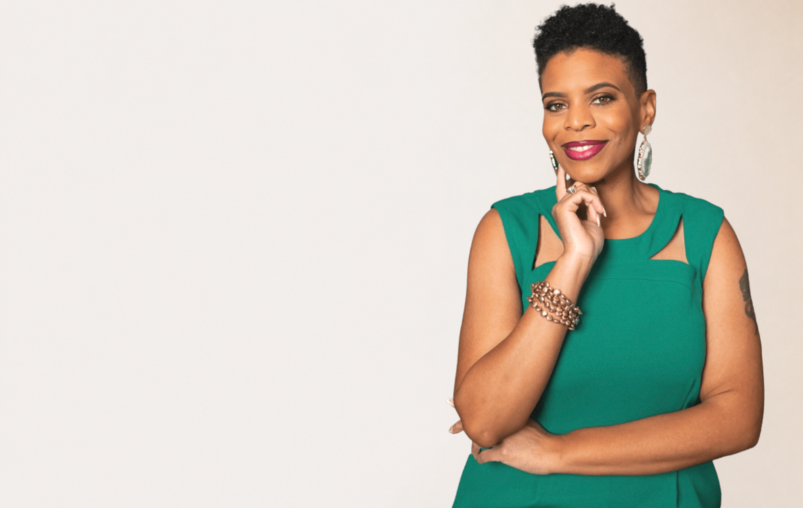 A woman with short curly black hair, wearing a sleeveless green dress with cutout details on the shoulders, smiling and posing with her hand resting on her chin, accessorized with large earrings and a bracelet, against a plain light background.