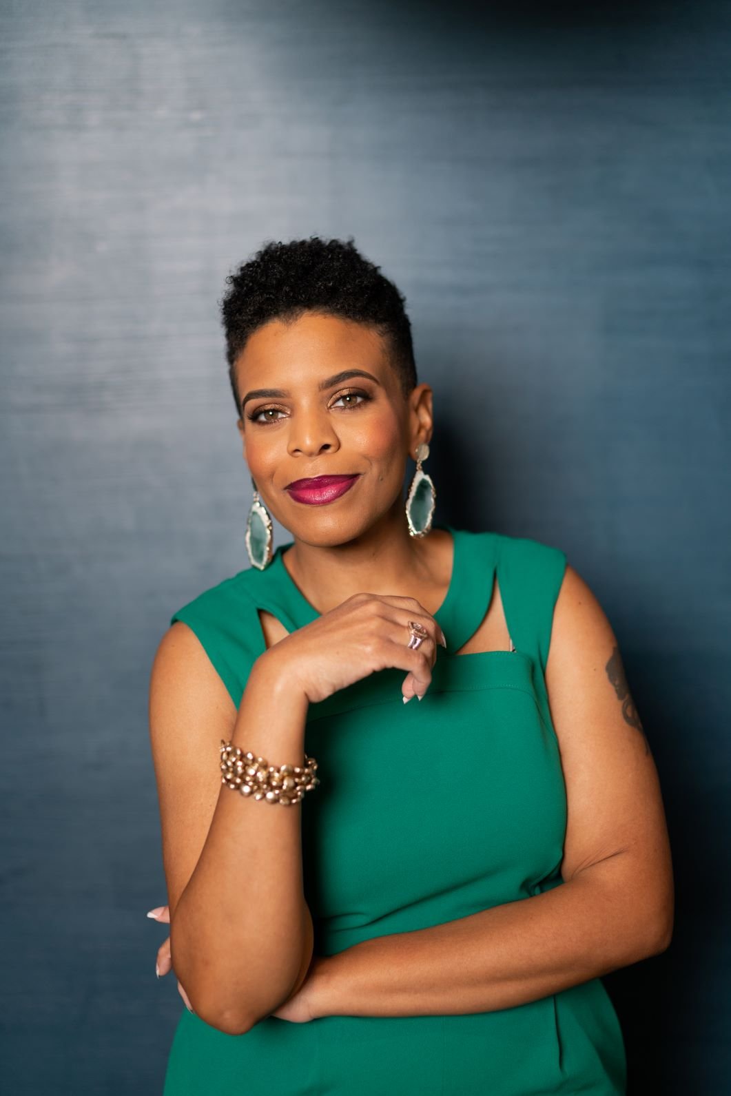 A woman with short curly hair and jewelry, wearing a green dress, stands in front of a blue background, looking confidently at the camera.