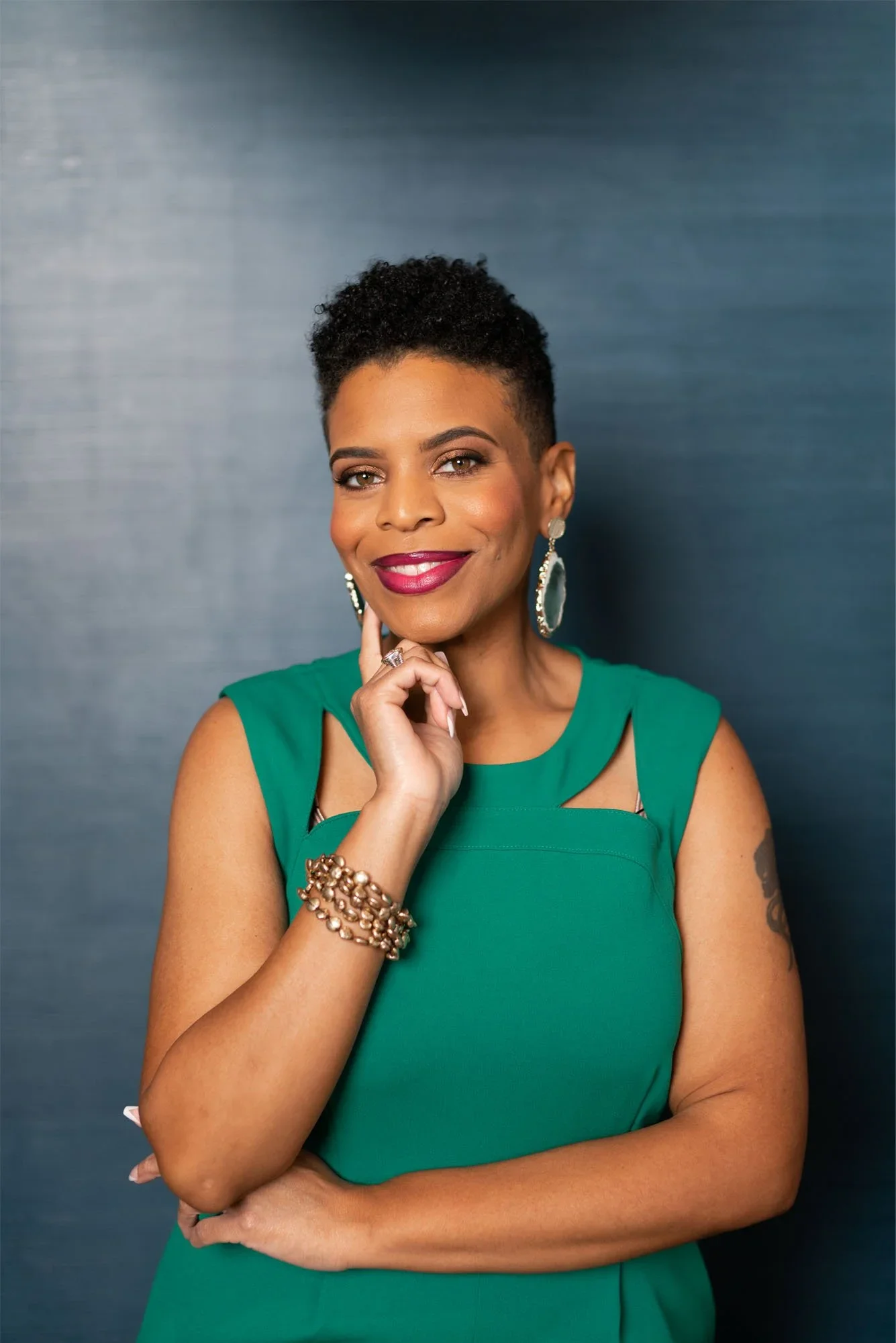 Stacey is pictured with short curly black hair, wearing green dress and large earrings, smiling with her hand on her chin, standing against a dark background.