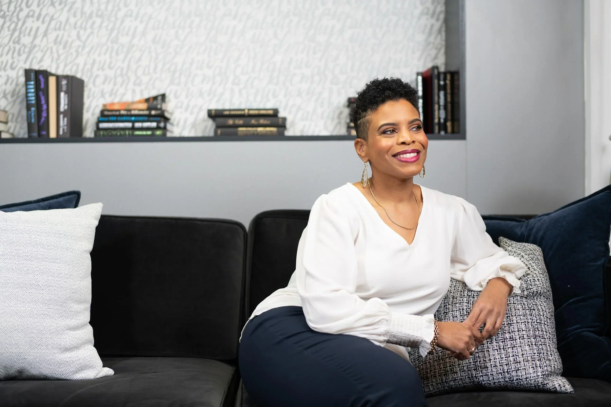 Stacey is pictured with short curly hair and jewelry sitting on a black sofa, smiling in a modern living room with bookshelves and decorative pillows.