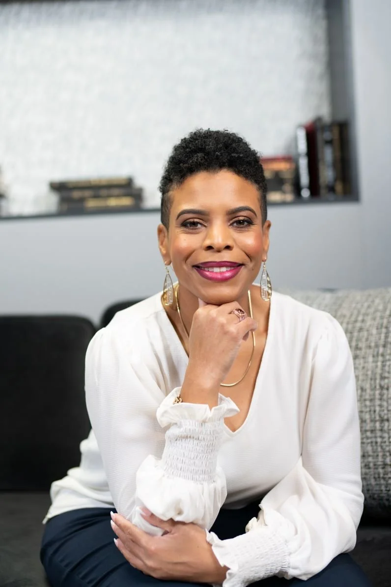 Stacey is pictured smiling with her fist under her chin with short curly hair, wearing a white blouse and jewelry, sitting on a black sofa in a modern room with a bookshelf in the background.