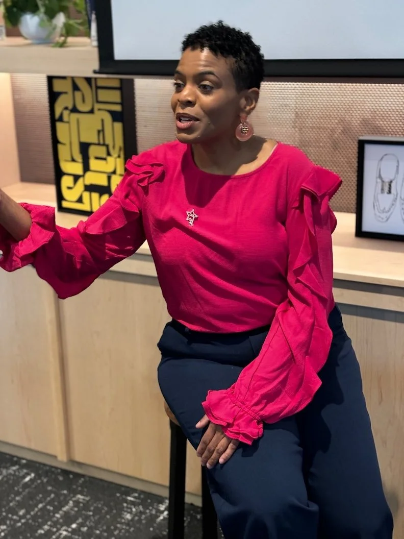 Stacey is pictured with short black curly hair wearing a bright pink blouse with ruffles, navy pants, and pink earrings, sitting on a stool and gesturing with her right hand in an indoor setting.