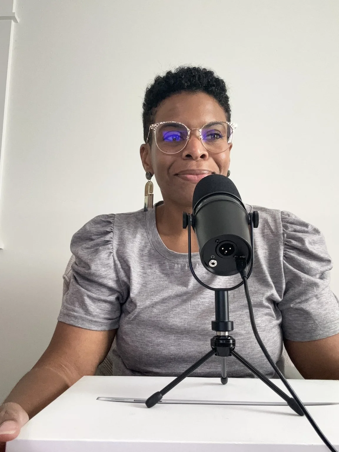 A woman with short curly hair, wearing glasses and earrings, is sitting at a table with a microphone in front of her. She is smiling and looking slightly to the side.