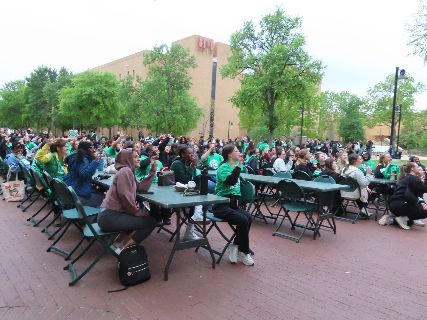 Large outdoor gathering of diverse students sitting at tables, some raising their hands, with a university campus building and green trees in the background.