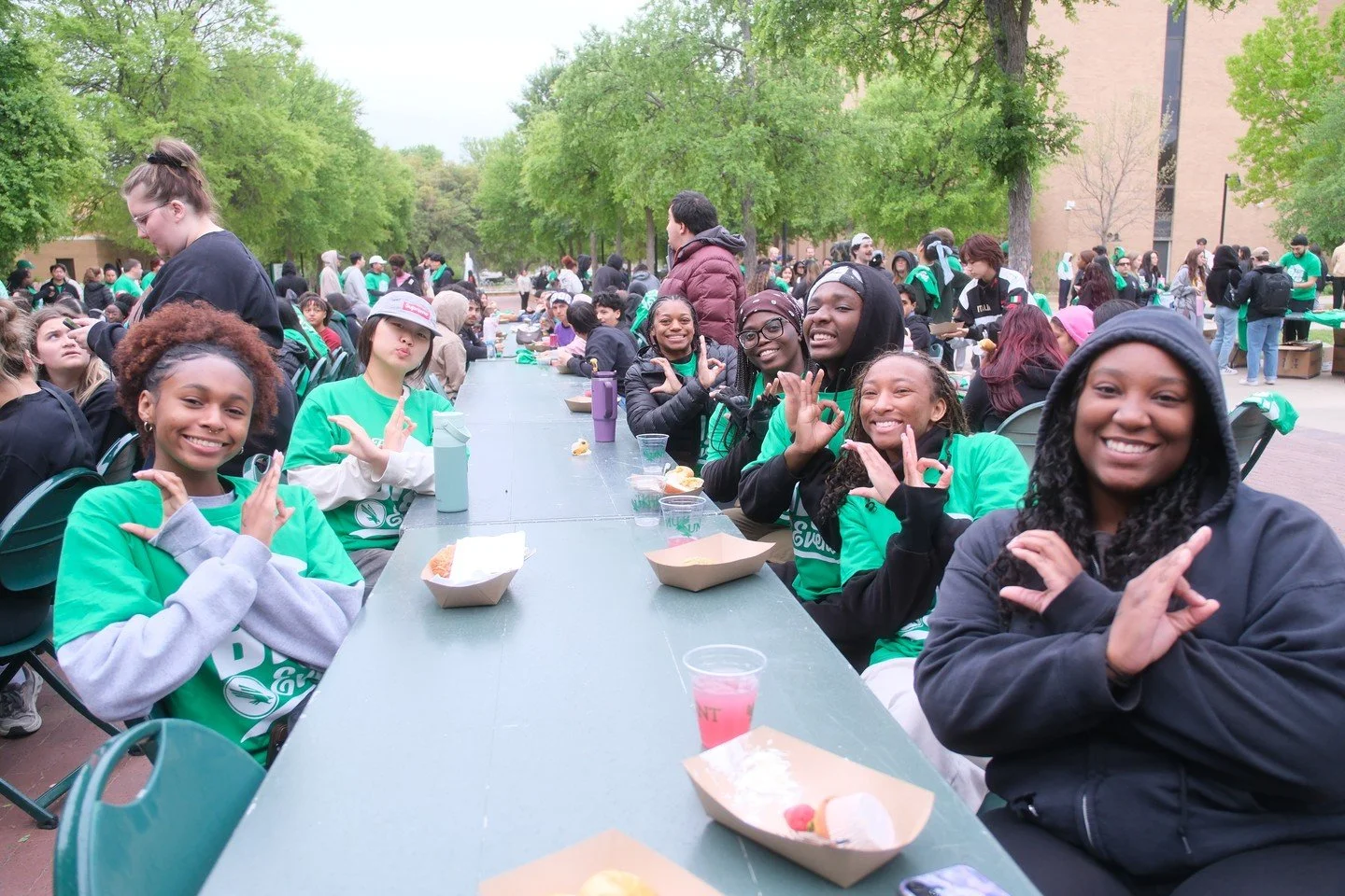 Group of young people sitting at a long outdoor table, smiling and making hand gestures, at a picnic or outdoor event, with trees and other people in the background.