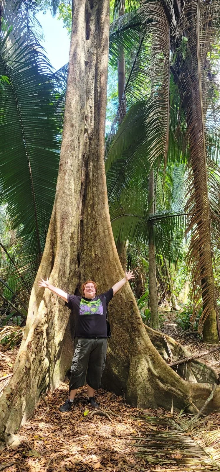 Person standing in a lush tropical forest, smiling with arms outstretched, in front of a large tree with roots spreading on the ground, surrounded by green palm and jungle leaves.