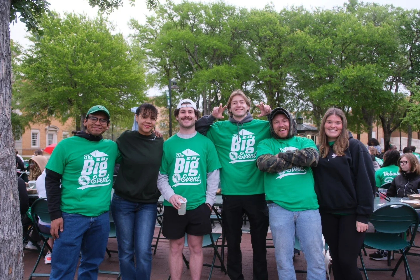 Six young adults standing together outside at an event, wearing green t-shirts with a logo that says 'The Big Event.' They are smiling and appear to be friends or volunteers. There are trees and other people in the background.