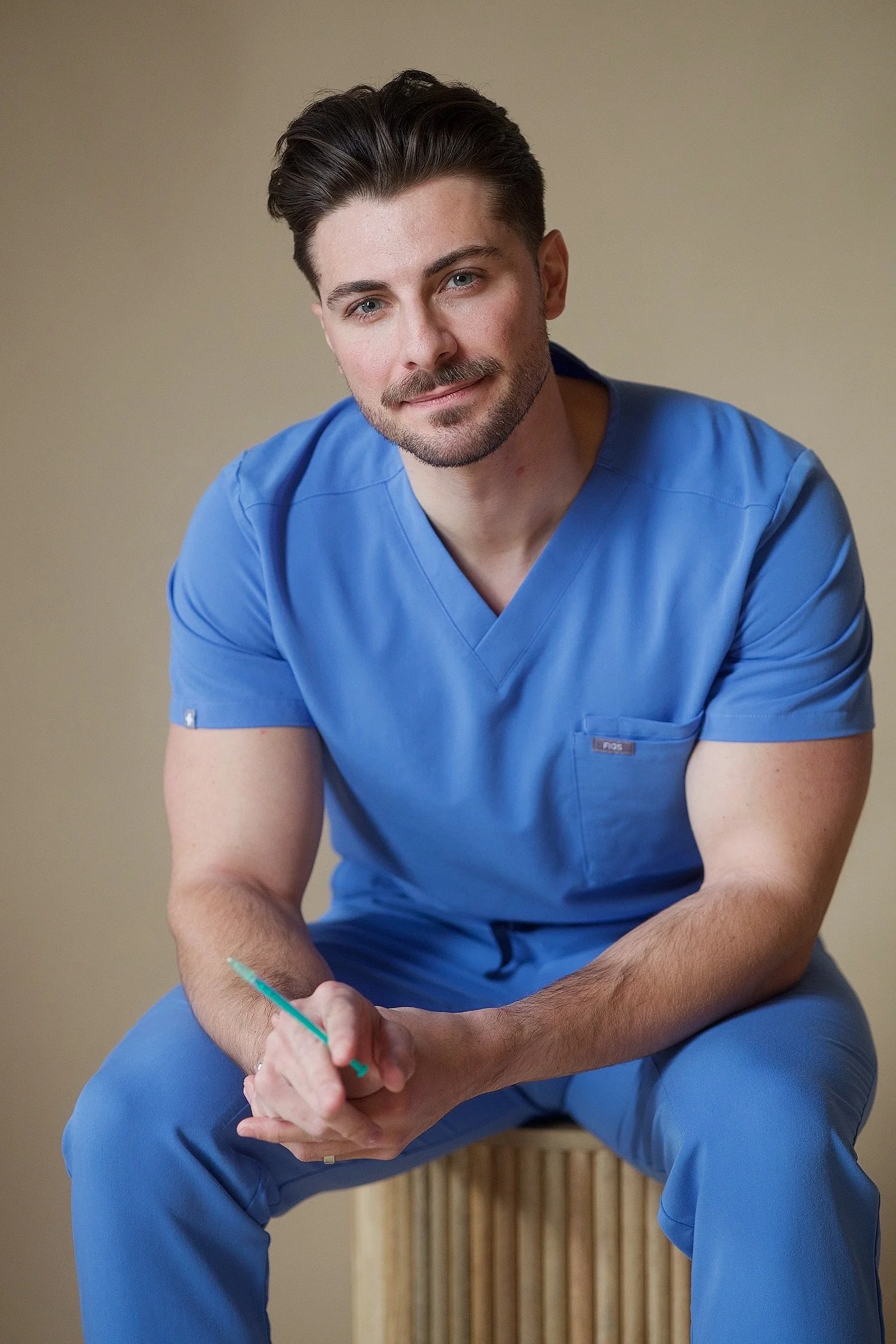 A young male healthcare professional in blue scrubs sitting against a beige background, holding a syringe.