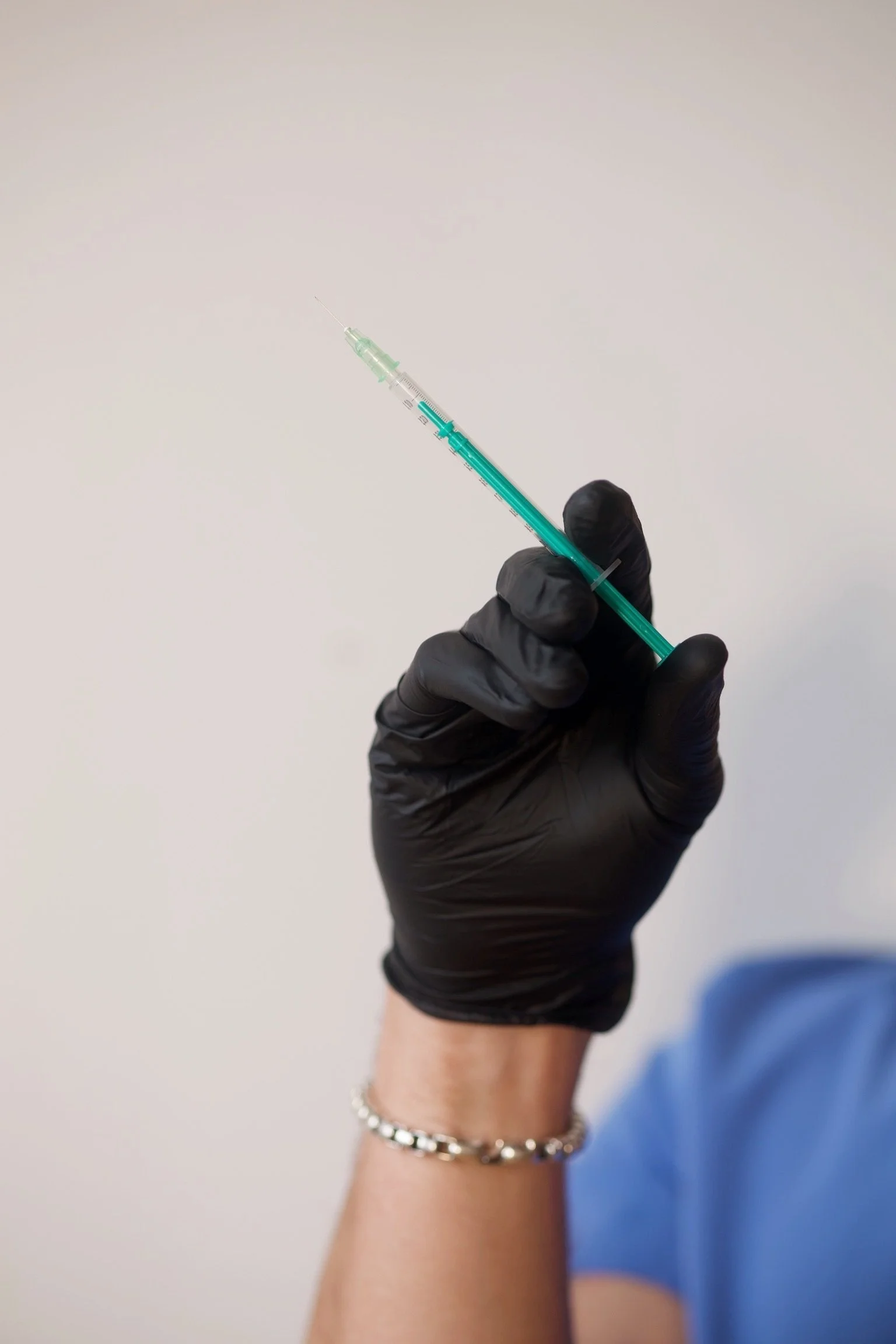 A hand in a black glove holding a syringe filled with a liquid, with a blurred arm and a blue shirt in the background.