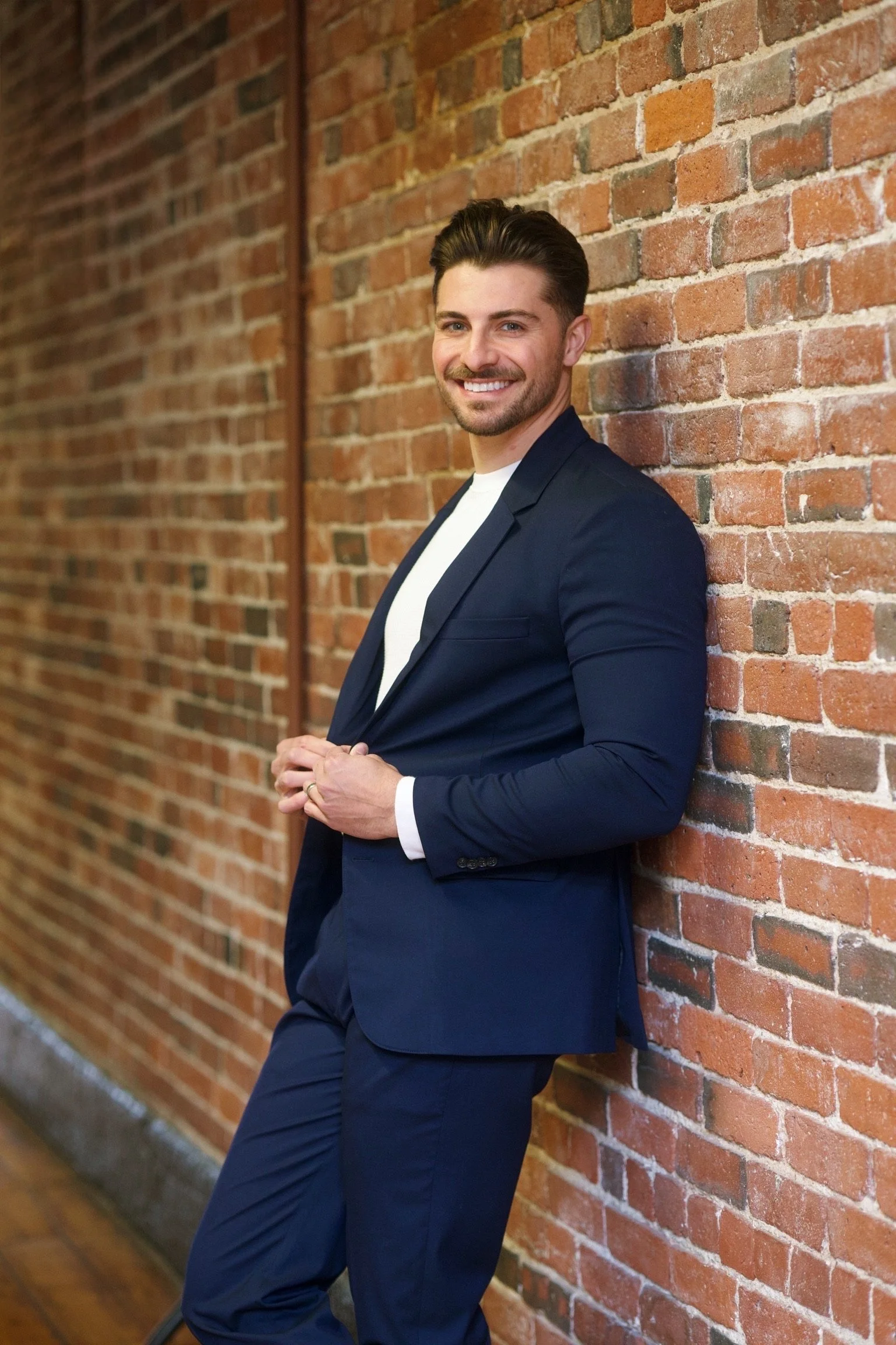 A young man with dark hair, a beard, and a bright smile, dressed in a navy blue suit and white shirt, leaning against a brick wall with a relaxed pose.