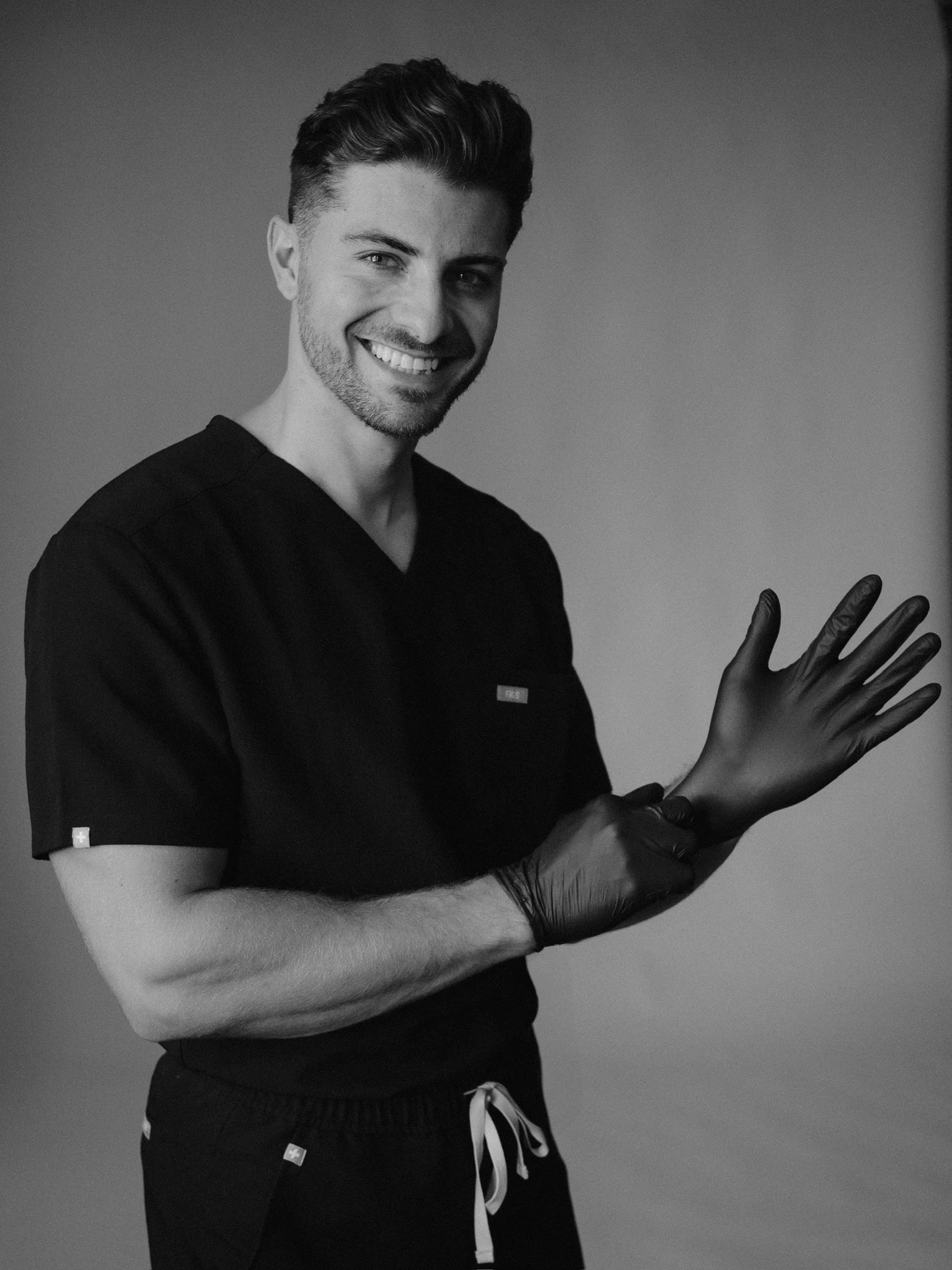 A smiling man with styled hair and a beard wearing a dark medical scrubs top and black gloves, holding his hand open in front of him, with a plain gray background.
