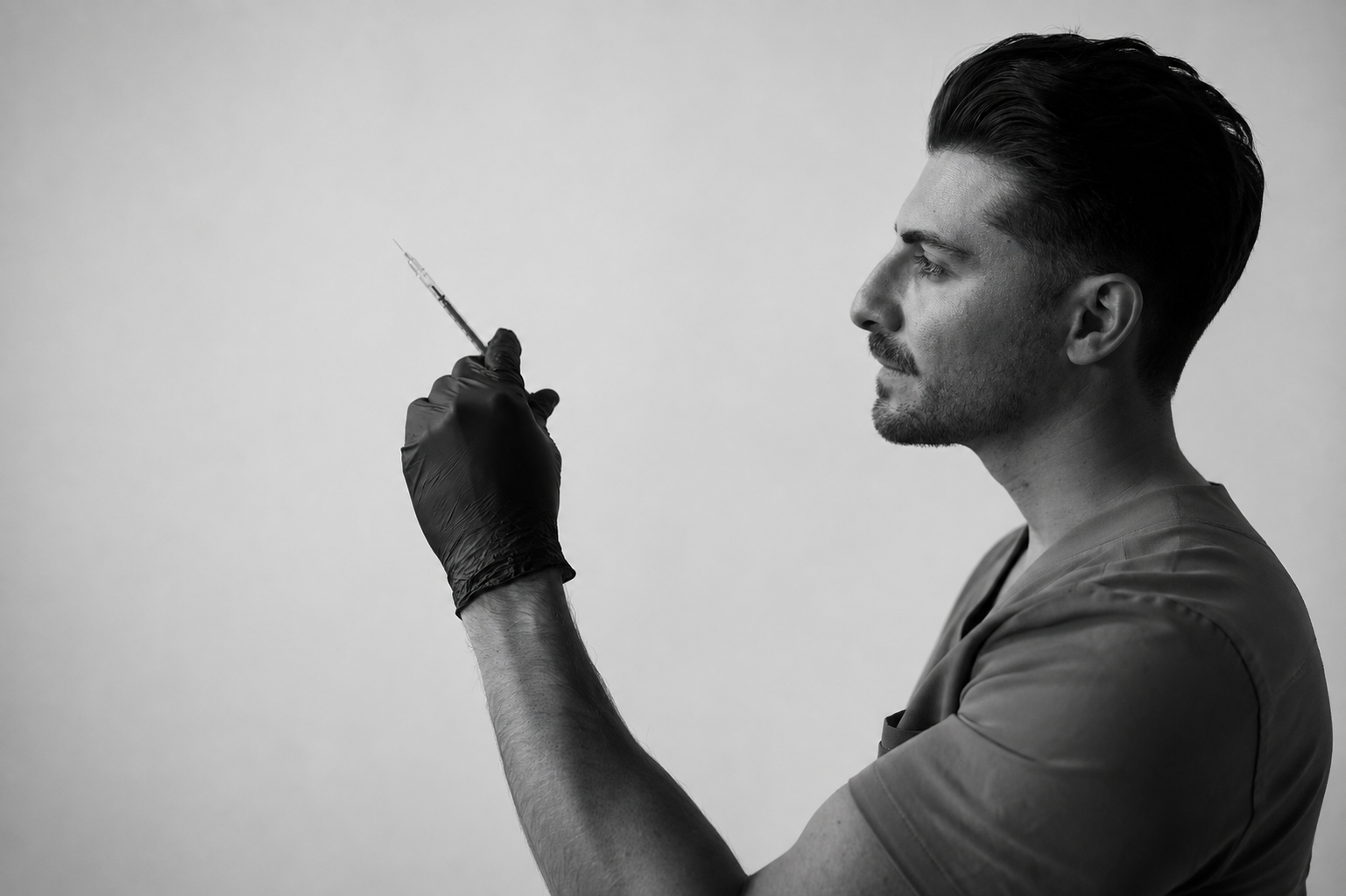 Black and white photo of a man in medical scrubs holding a syringe while looking at it thoughtfully.