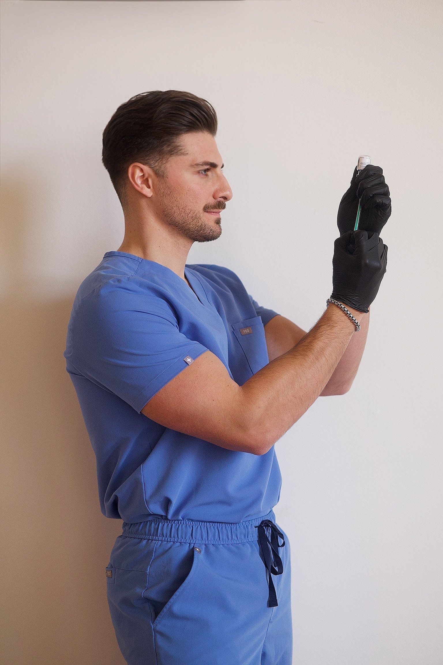 A young man in blue medical scrubs with a name tag, black gloves, and a bracelet, holding a test tube and looking at it intently.