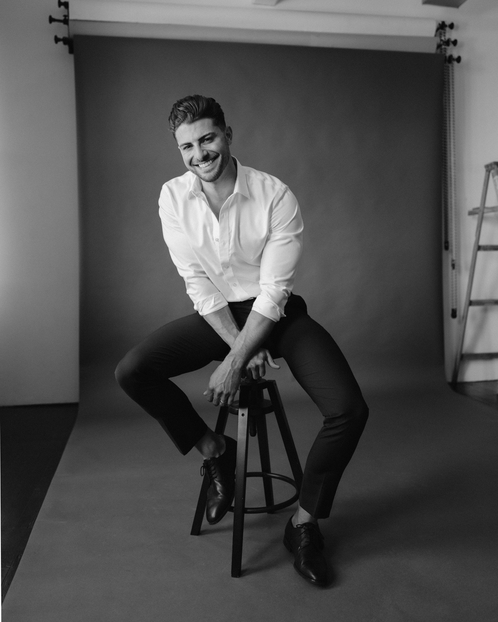 Black and white photo of a man sitting on a chair, smiling, wearing a white shirt and black pants, with one leg crossed over the other, in a photography studio with a backdrop and a ladder to the side.