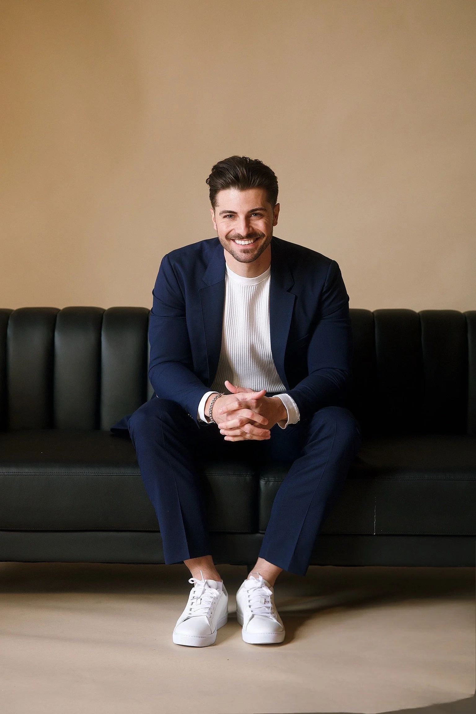 A young man with dark hair and a beard, smiling, sitting on a black leather sofa against a beige background, wearing a navy blazer, white shirt, navy pants, and white sneakers.