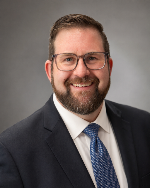 Headshot of a smiling man with glasses, a beard, and short brown hair, dressed in a dark suit, white shirt, and blue tie, against a gray background.