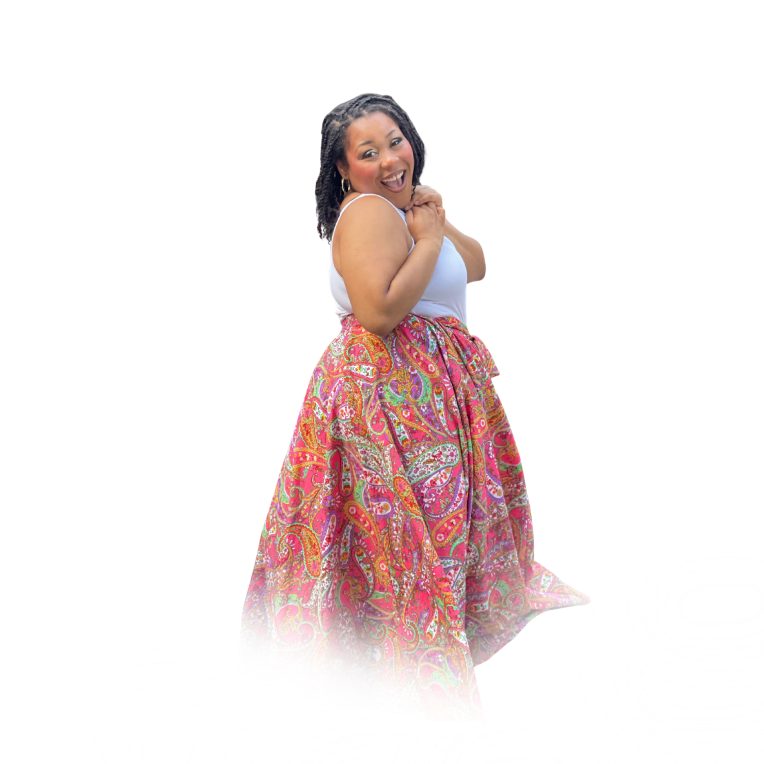 A joyful woman with dark curly hair wearing a white tank top and a colorful, patterned skirt, standing with her hands near her chest, smiling brightly.