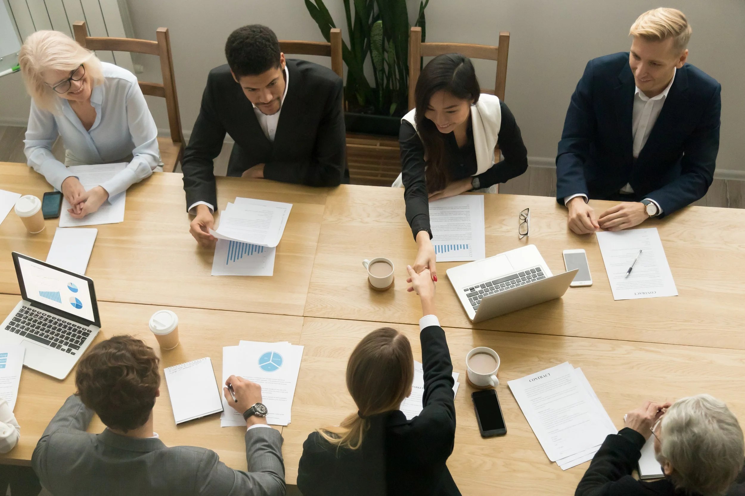 Group of professionals sitting around a meeting table, discussing documents, with laptops, smartphones, and coffee cups present.