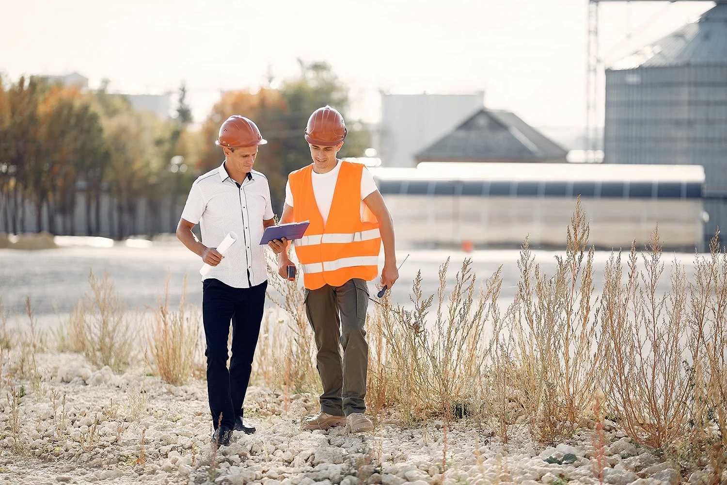 Two construction workers walking on a gravel site, wearing safety helmets. One in an orange safety vest is holding a tool and a clipboard, while the other is holding a rolled-up paper and looking at the clipboard, discussing plans. Background shows construction site with buildings and trees.