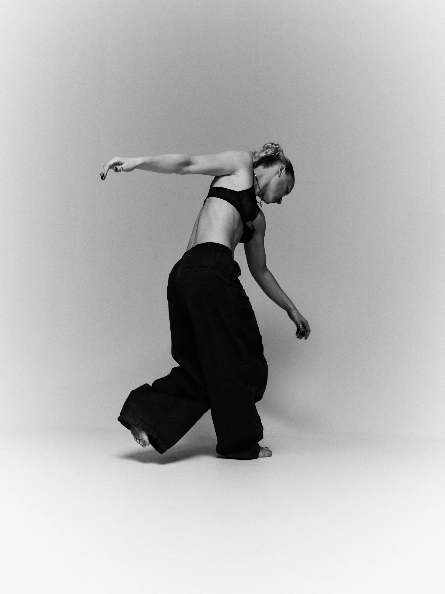 A person with a shaved head and glasses performs a dance move, leaning forward with one arm extended out to the side and the other arm hanging downward, wearing a black crop top and loose black pants, against a plain light gray background.