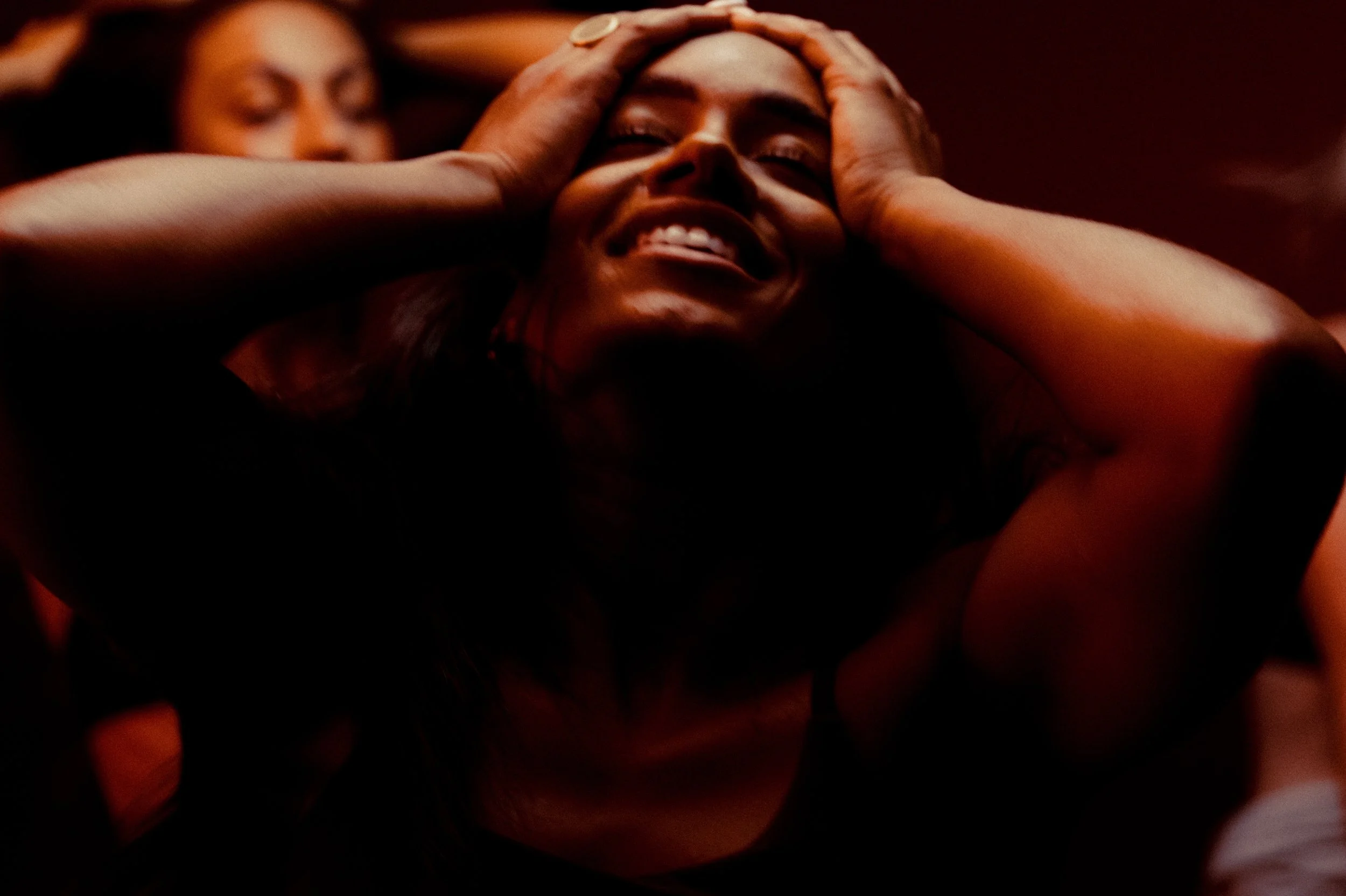 A woman smiling and holding her head with both hands, with another woman in the background and a dark, warm lighting setting.
