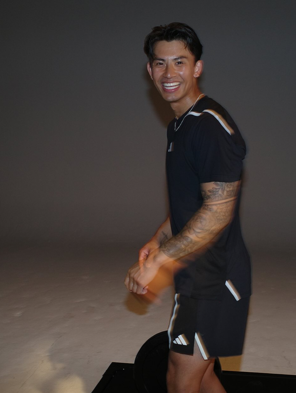 A smiling man with tattoos on his arms, wearing a black sports outfit with white accents, standing indoors against a dark background.