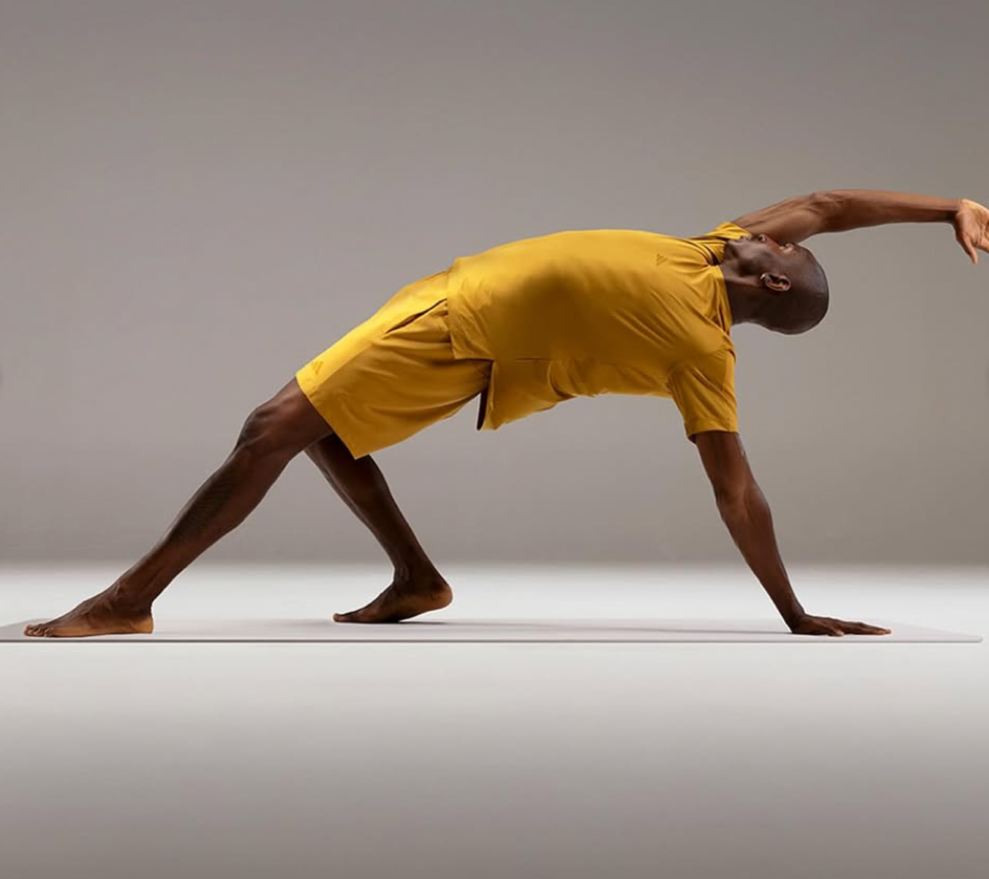 A man in a yellow athletic outfit is doing a yoga pose on a white mat in a studio with a plain gray background.